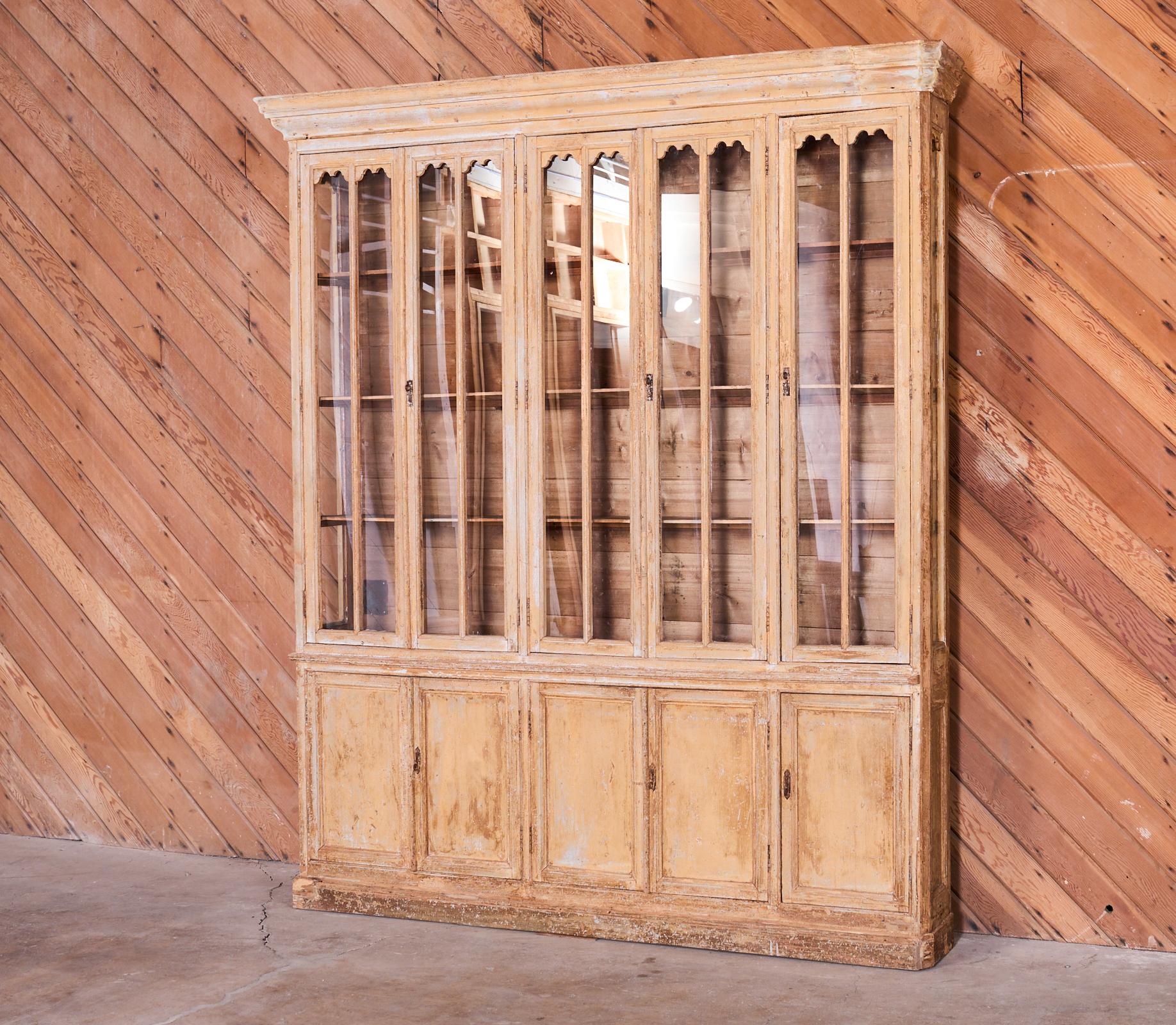 Rustic and weathered 19th century country French provincial library bookcase cabinet with glass doors. The large case features a stripped wood finish with old paint remnants. The wood has a bleached aged patina on the finish. The cabinet is one