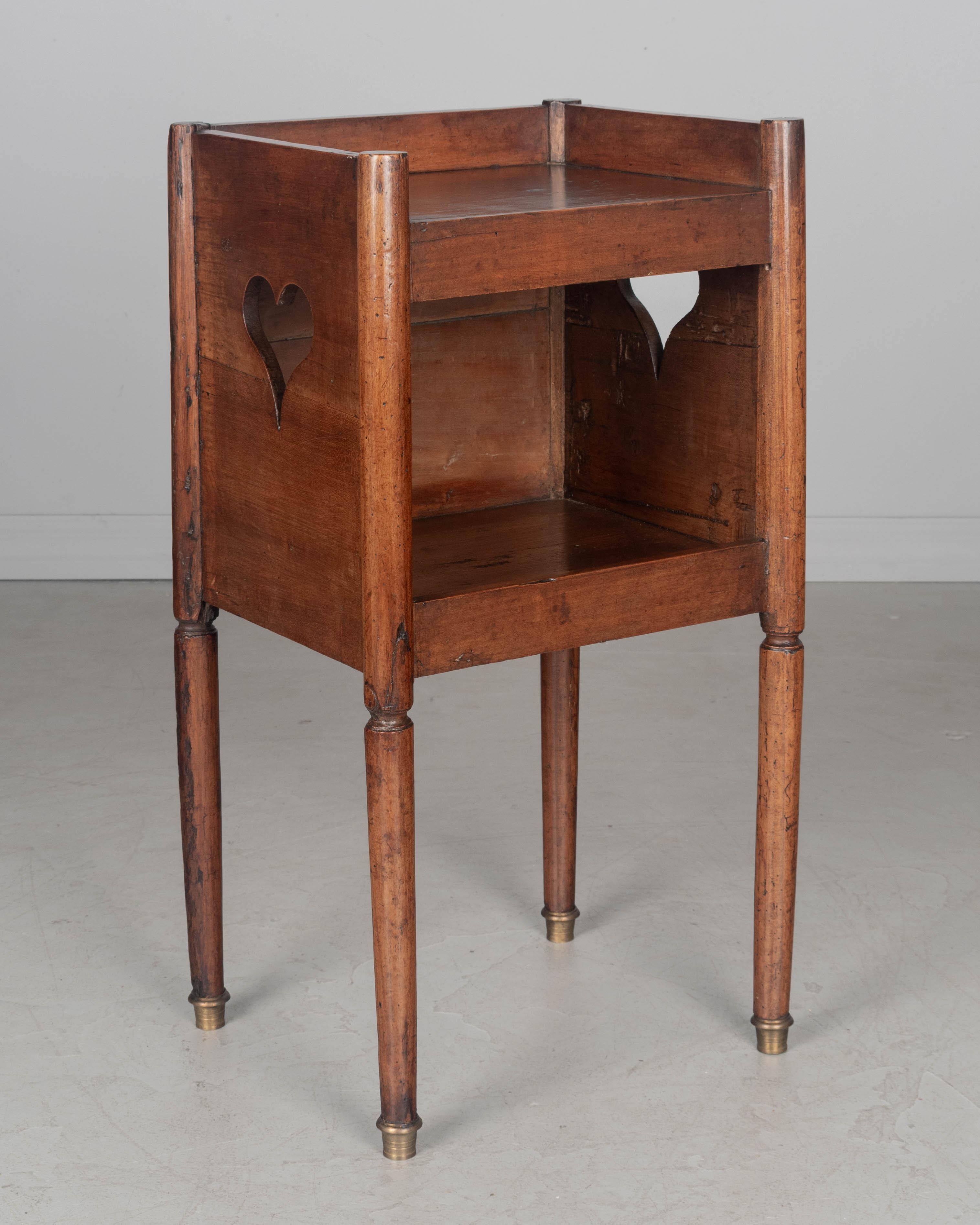 Table d'appoint ou table de nuit de style campagnard français du XIXe siècle, en noyer massif, avec des découpes en forme de cœur sur les côtés et une niche ouverte. Pieds fuselés tournés avec sabots en laiton. Finition sur les quatre côtés. Une