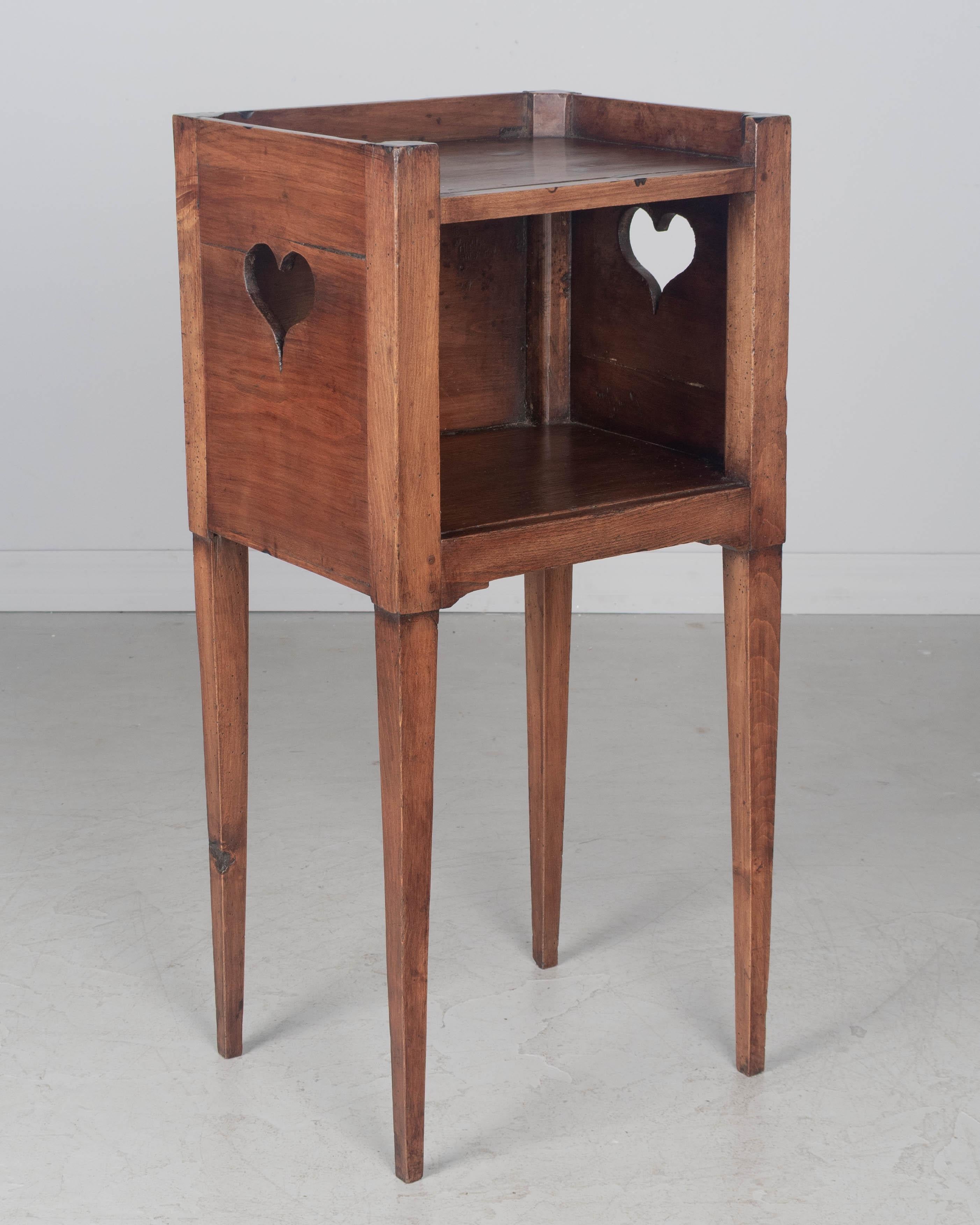 Table d'appoint, ou table de nuit, de style campagnard français du XIXe siècle, avec des découpes en forme de cœur sur les côtés et une niche ouverte, et de minces pieds fuselés. Fabriqué en bois de noyer, de cerisier et de hêtre et fini sur les