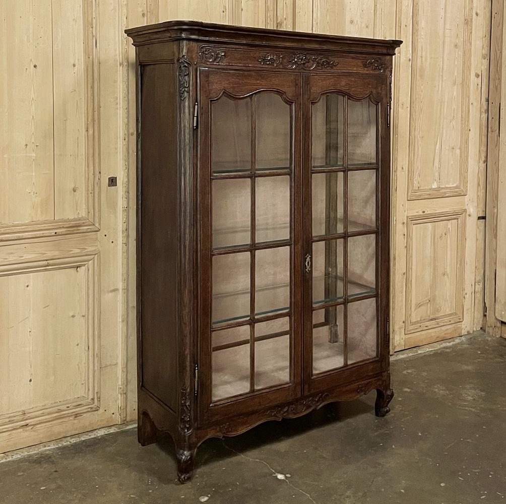 19th century country French vitrine, bookcase is the ideal choice for a cozy office or study! Hand-crafted from dense, old-growth indigenous oak, it has been masterfully carved with a floral basket flanked with shell and foliate sprays on the
