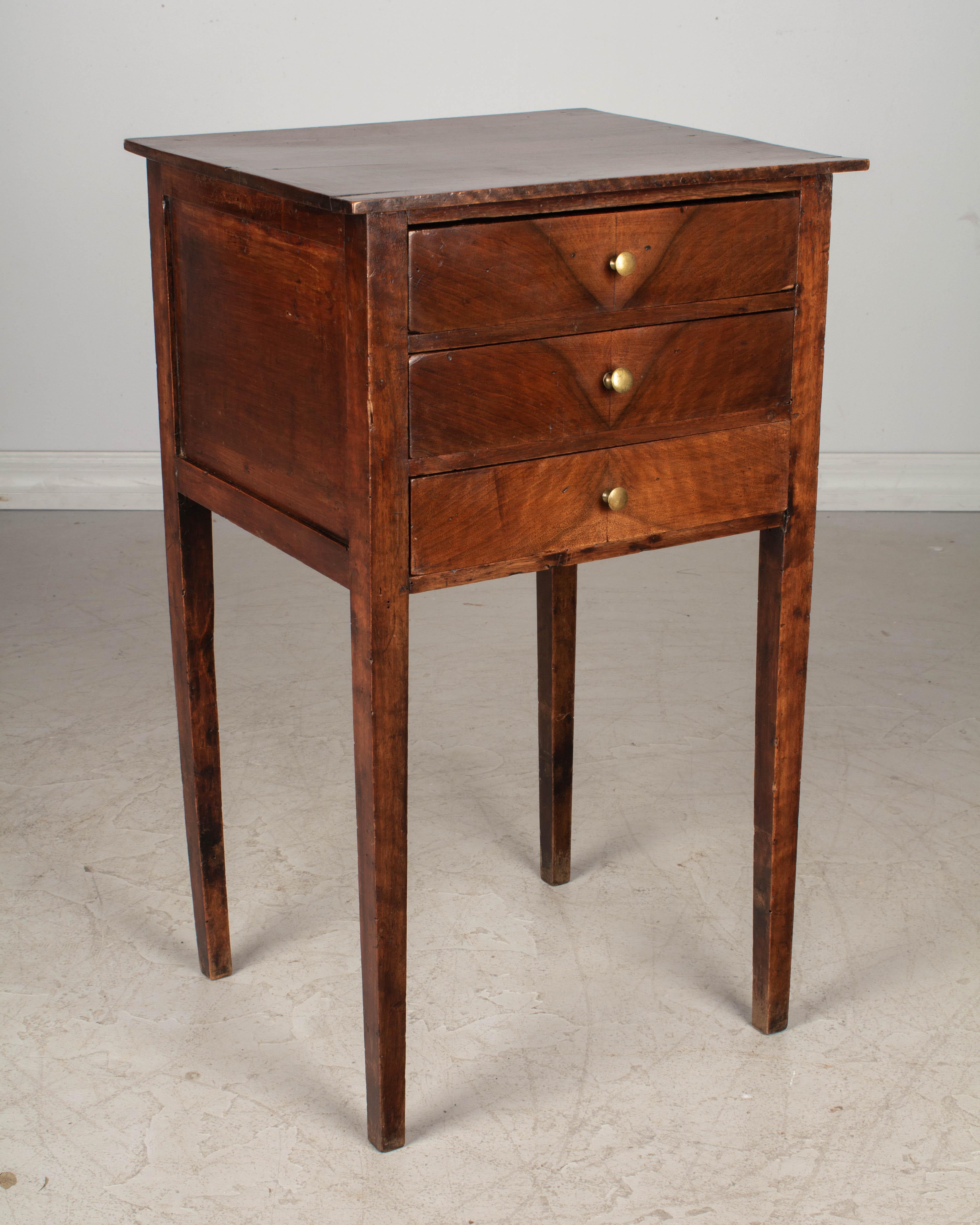 A late 19th century Country French walnut side table, or nightstand, with tapered legs and three dovetailed drawers with book matched face and small brass knobs. Handcrafted of solid walnut using pegged construction and mortise and tenon joints.
