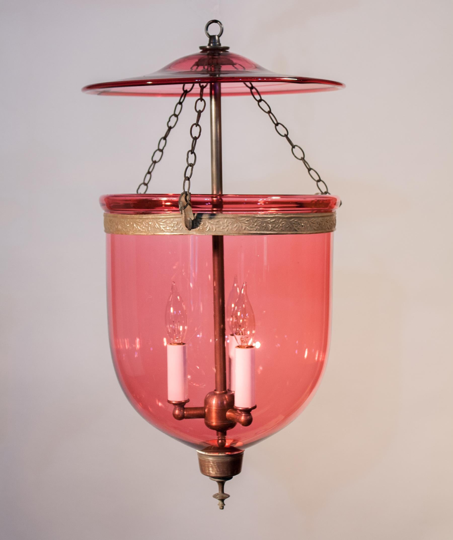 An exceptional cranberry-colored hand blown glass bell jar lantern from England. Adorned with its original brass band and finial base, this pendant light casts a warm, beautiful light. The light fixture has been newly electrified with a three-bulb