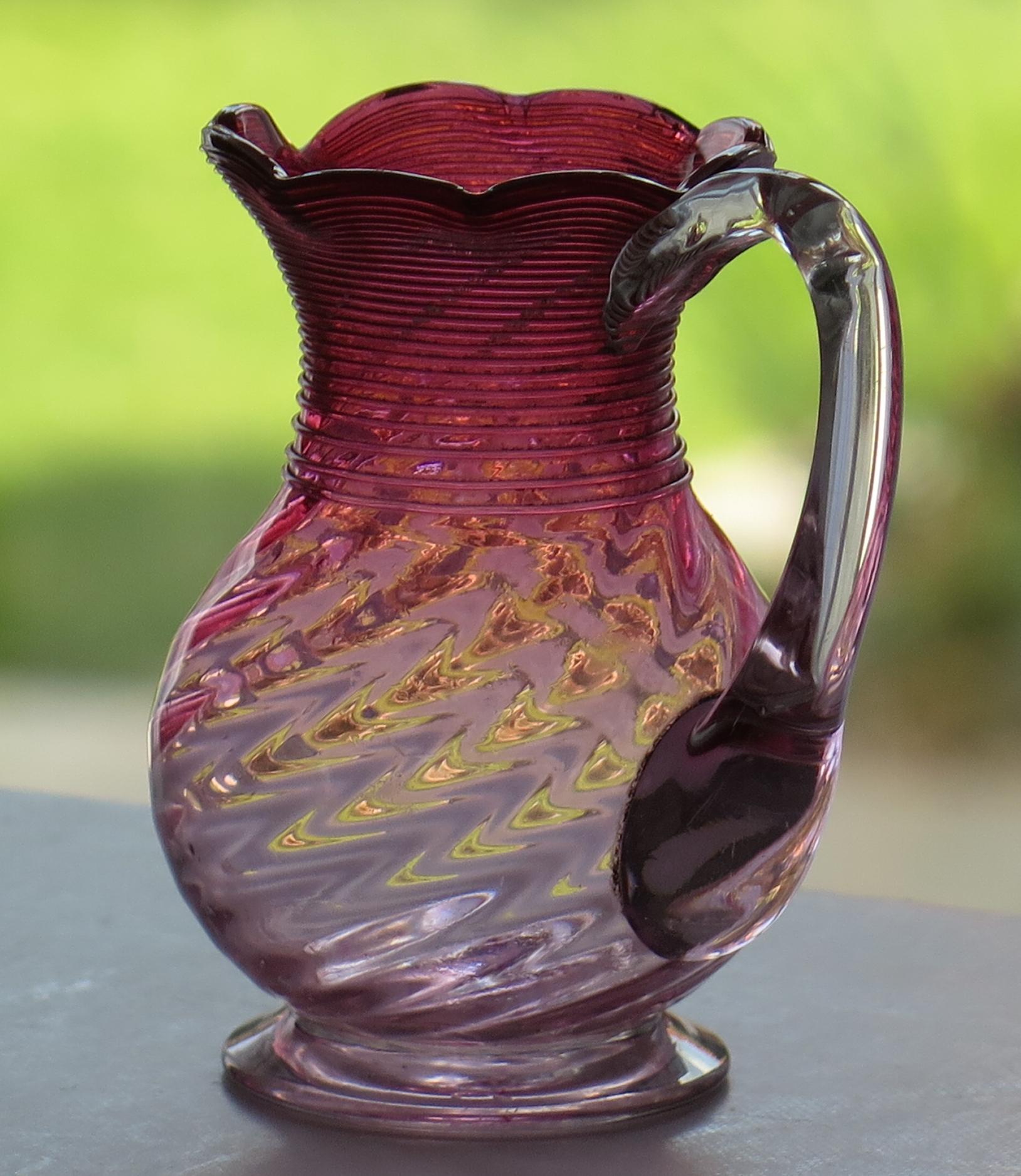 This is a good wrythen (spiral) moulded cranberry glass milk or cream jug, which we date to the mid-late 19th century of Victorian England, circa 1870.

The body is made from cranberry glass with an applied clear glass loop handle

The jug has a