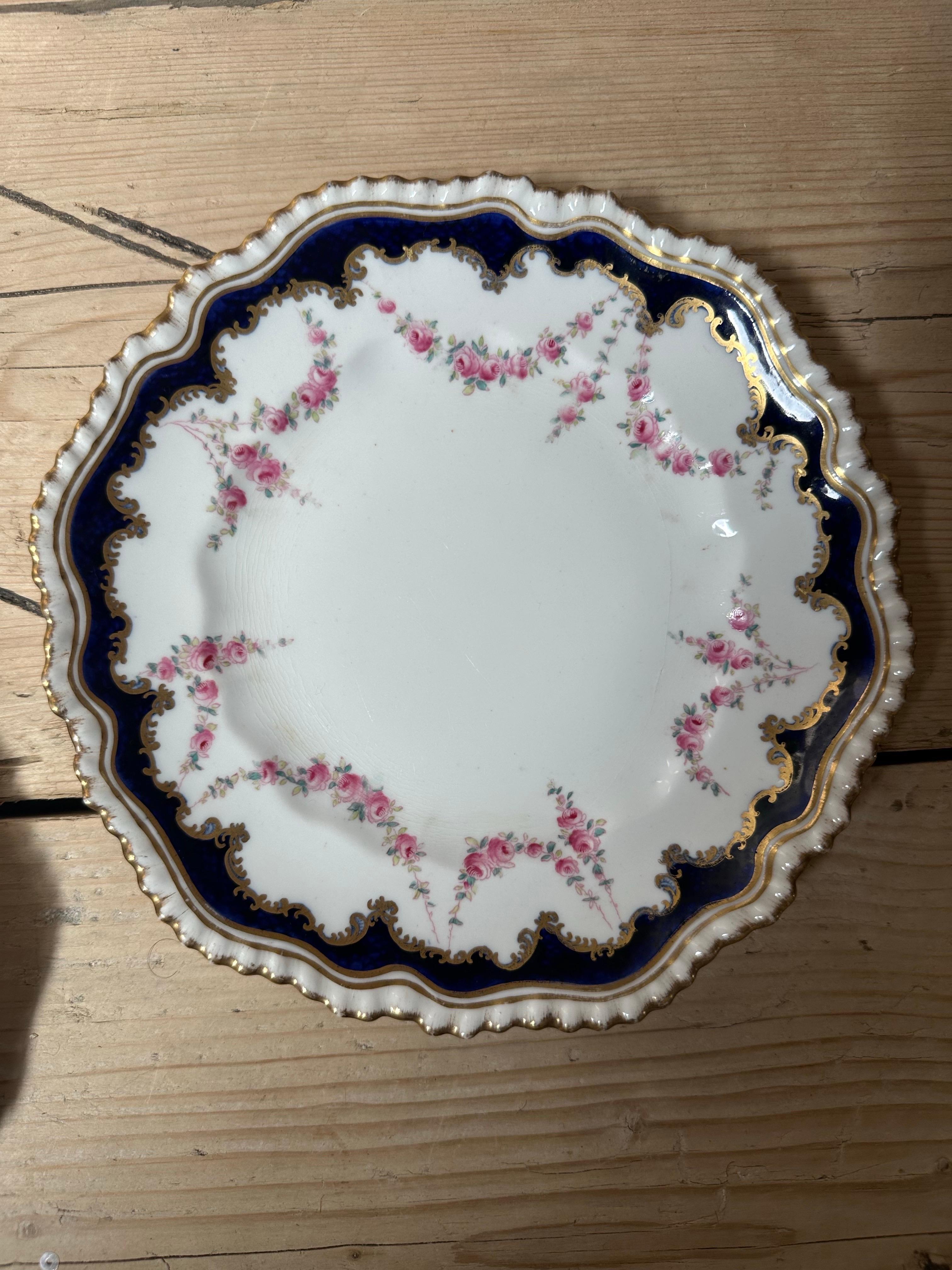 The image shows an antique porcelain plate, likely a dessert or serving plate, featuring a distinctive design with a dark blue, scalloped rim and a delicate rose garland motif. 

 The plate is from a prominent English porcelain manufacturer Crown