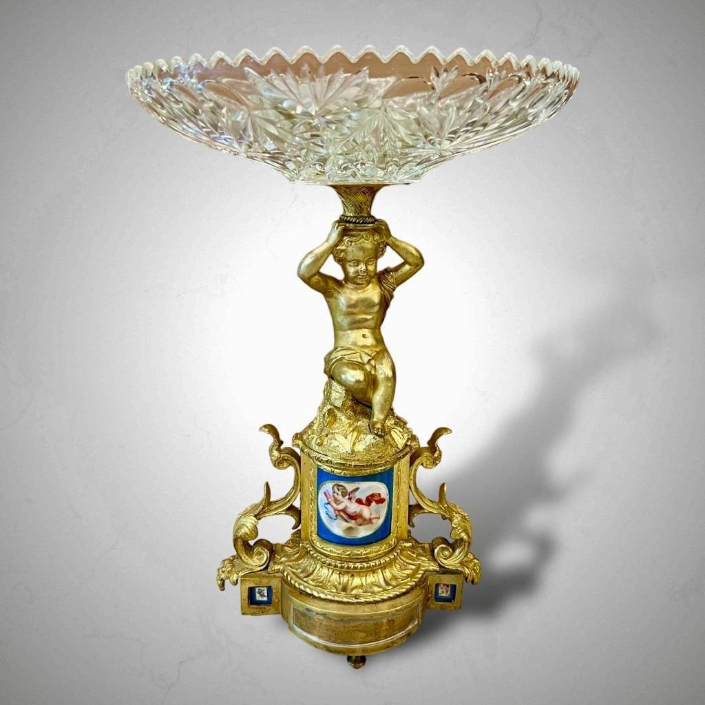We present this cut crystal bowl, elegantly poised on a gilt bronze pedestal adorned with a charming, seated putto, serving as a stem and holding the bowl on top his head. The base features two gilt bronze scrolls and is embellished with a porcelain