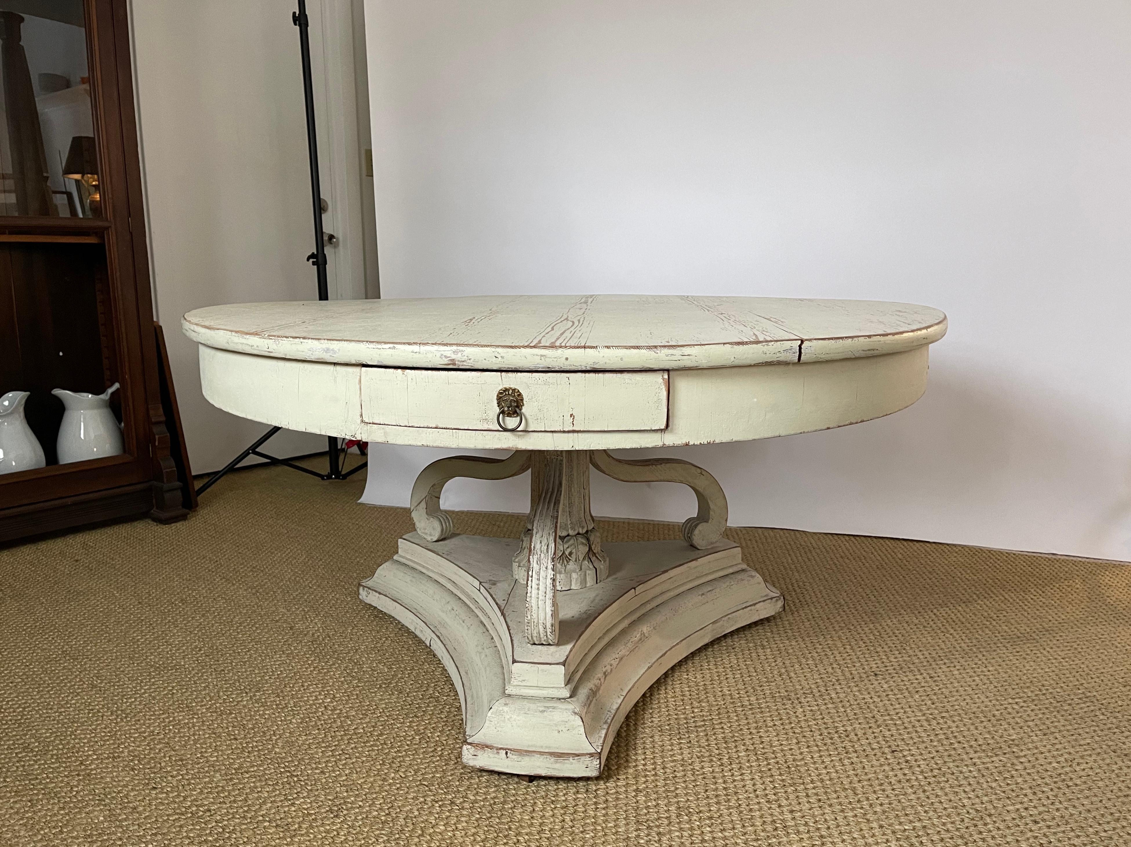 A 19th century Gustavian style Danish neoclassical white painted round pedestal dining or center table with three frieze drawers with brass lion mask pulls, supported by a fluted columnar base with carved foliate decoration and three scrolled