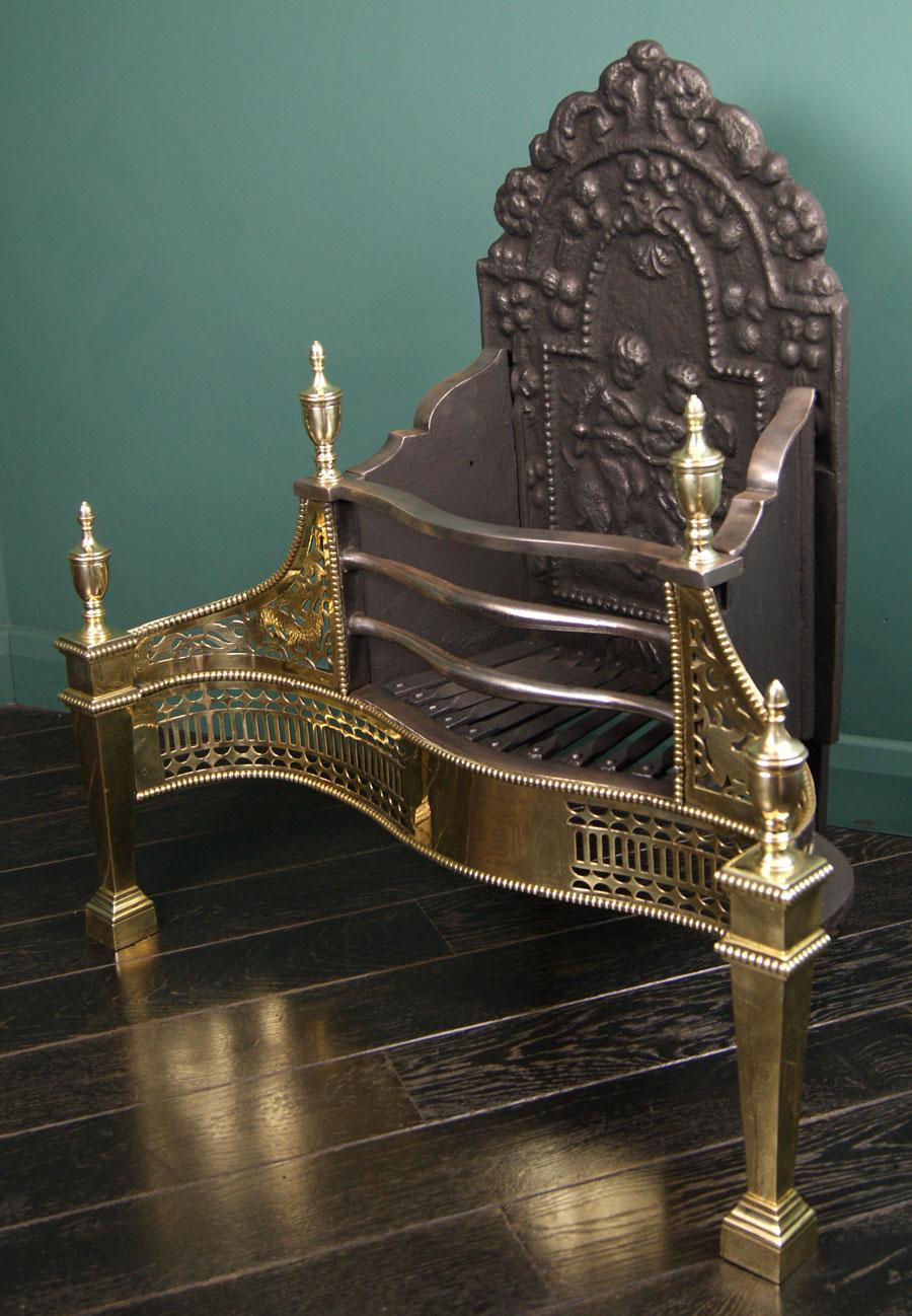 A 19th century decorative serpentine brass and steel dog grate.  The railed basket flanked by engraved dragon pierced wings edged with beading are set over a pierced fret and square tapered column legs.  Urn finials uppermost. Rear elements include