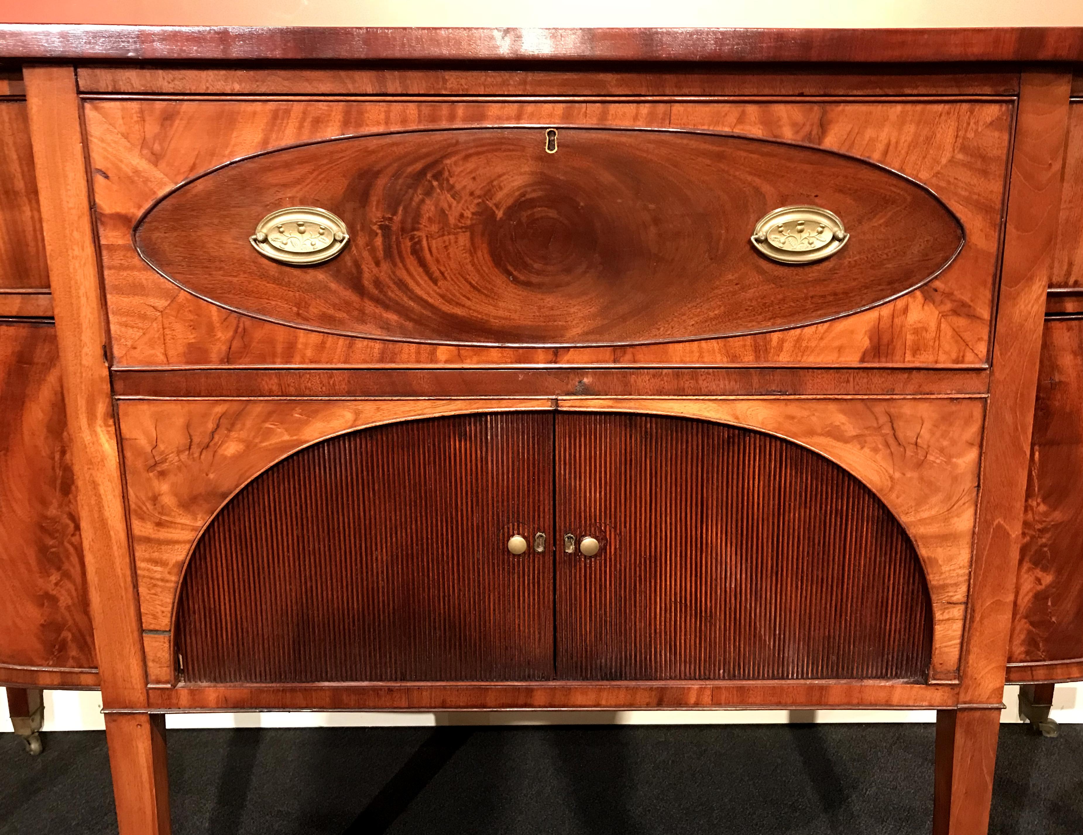 An exceptional Hepplewhite demilune form mahogany sideboard with conforming top surmounting a center pull-out butler’s desk which opens to a compartmentalized interior and writing surface, over two tambour doors which slide open to reveal a storage