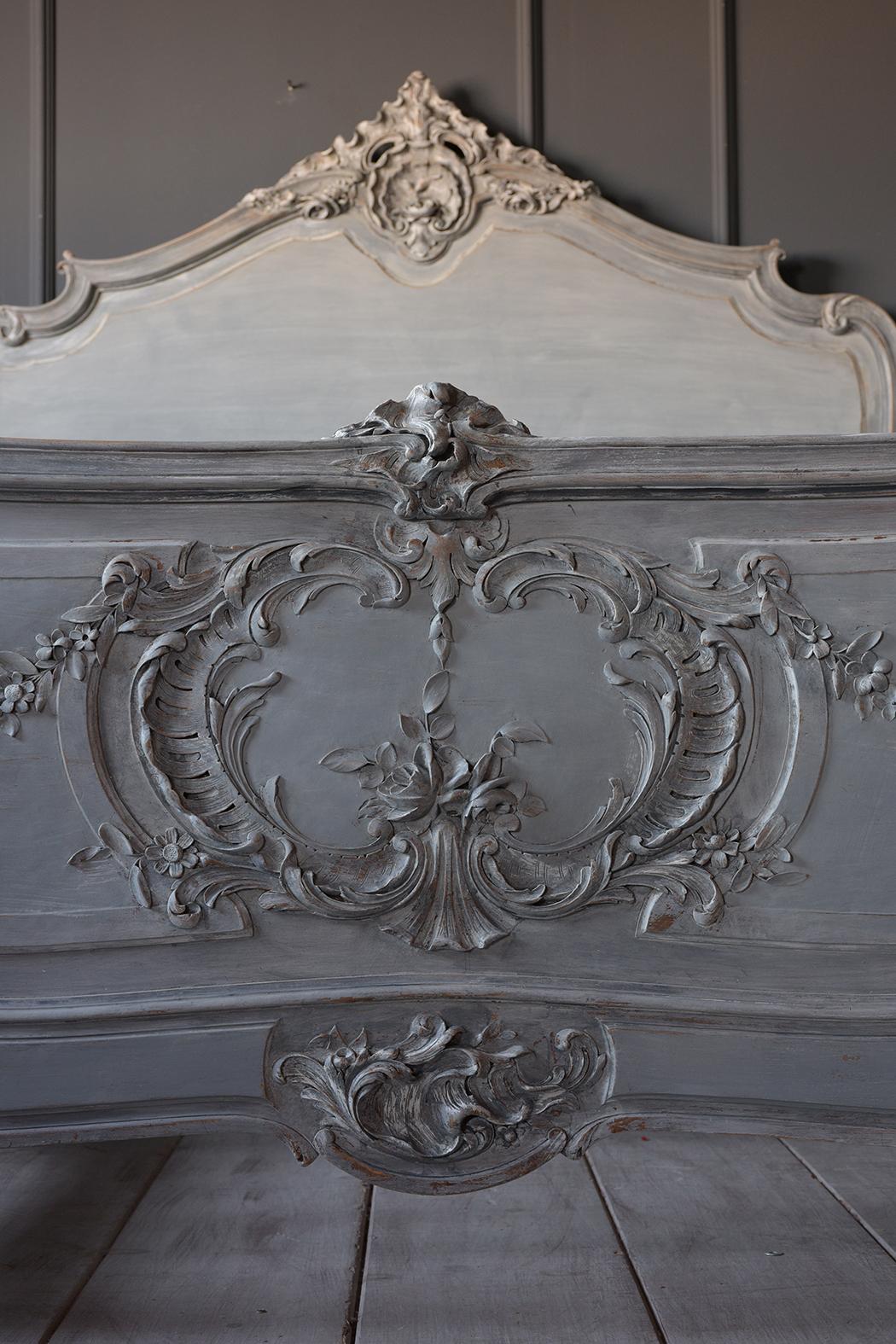 This French 19th century Louis XV style bed frame fits a full-size mattress. It has a solid wood frame that is painted a pale light blue, and grey with a distressed finish. It has carved details on the headboard, footboard, and wooden rails. The