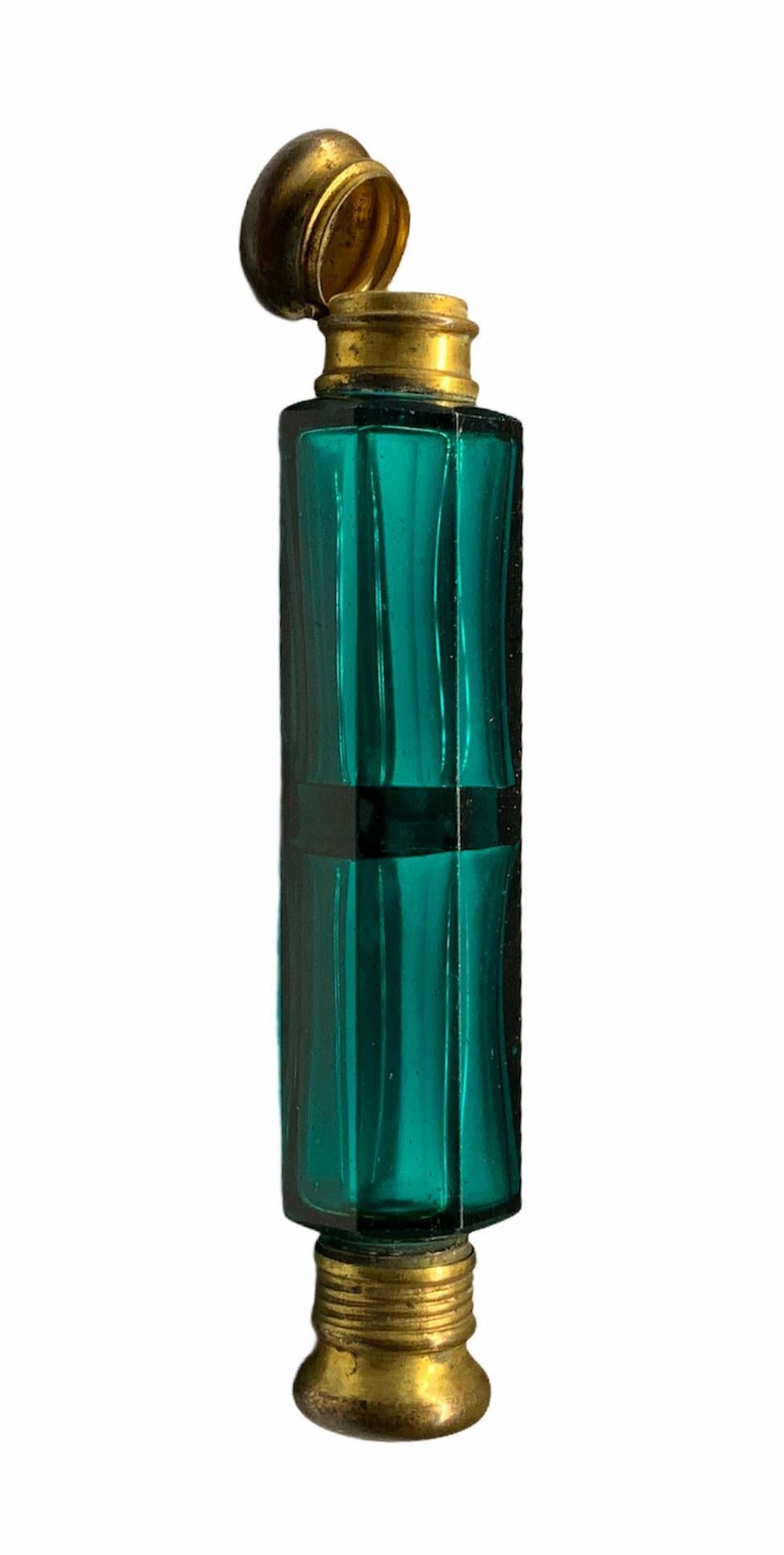 This is a double scent green glass long bottle with brass hinged cap with glass stopper in one side, and screw cap in the other end without stopper. The bottle is cylindrical shaped and the glass is faceted.
