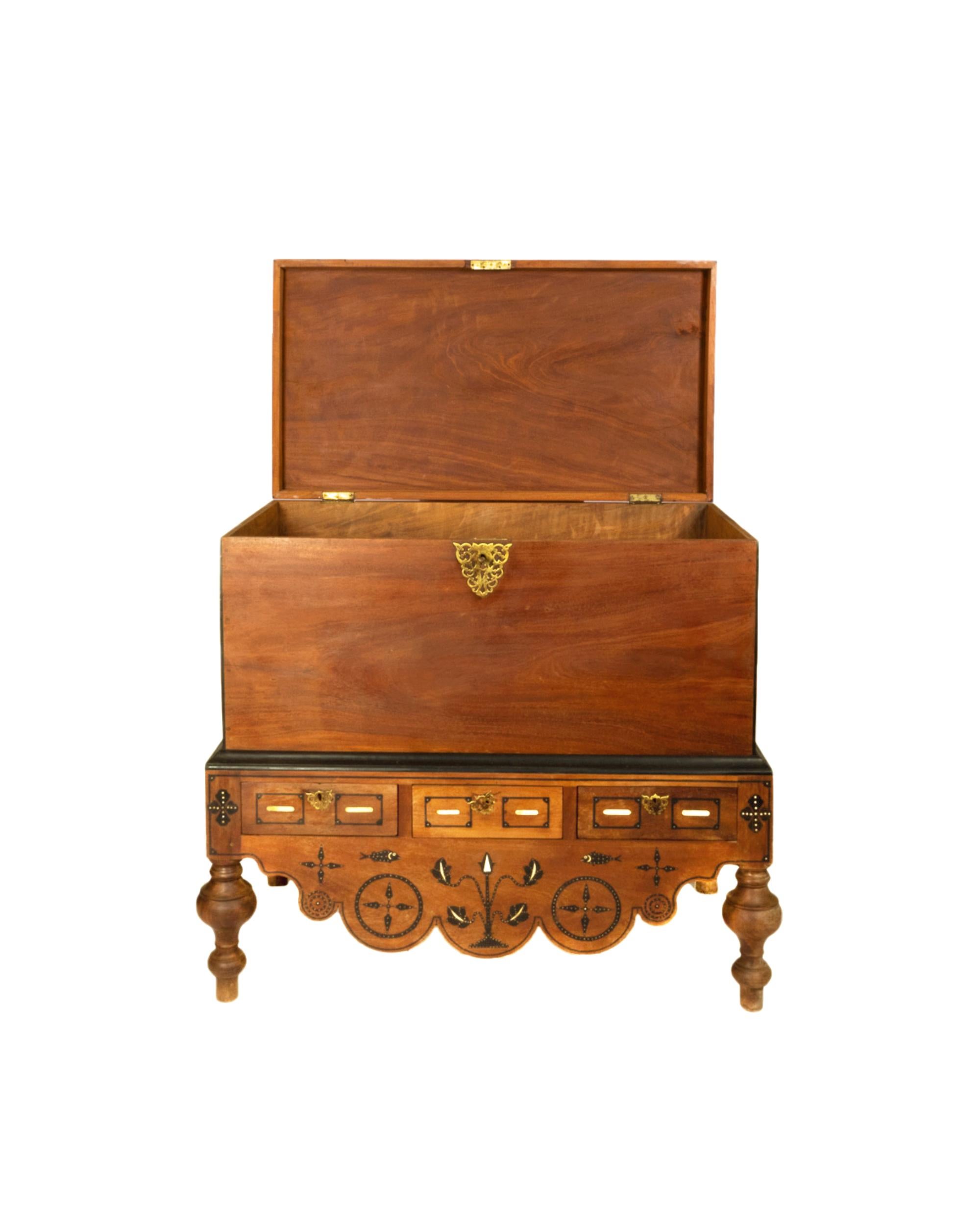 A stately Dutch Colonial chest in East Indian Satinwood, with hand turned legs, ebony trim, inlay, and brass accents and highly crafted marquetry in the lower section of the chest and lateral brass handles.  
The front has a deep, carved scalloped