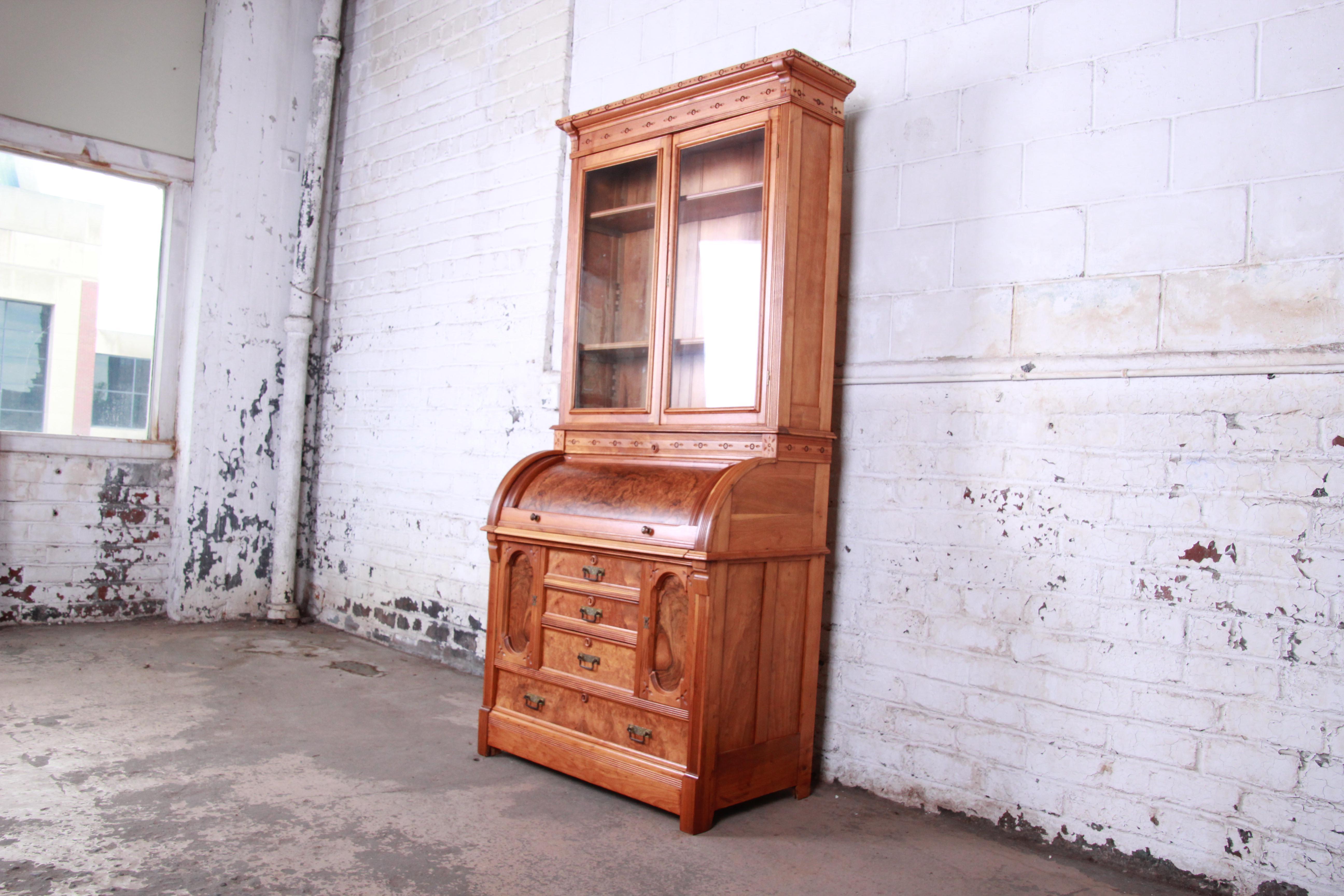 An exceptional 19th century Eastlake Victorian carved walnut and burl wood cylinder secretary desk with glass front bookcase. The desk features gorgeous burl wood grain and unique carved wood details. It offers ample storage and display. The top