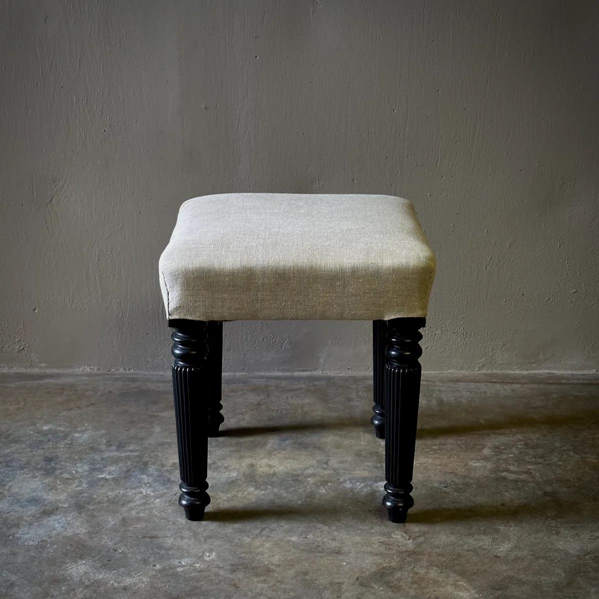 This late 19th-century English stool features fluted and turned ebonized wood legs, expertly reupholstered in flax-toned linen for a contemporary refresh. The piece’s modest yet elegant proportions make it a timeless addition to any room, offering