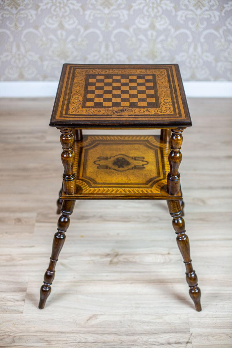 We present you a small oak table with a square top and a shelf.
This piece of furniture is dated 1897.
The shelf and top are covered with relief patterns in a darker shade of brown.
There is the motif of chessboard on the top and a date on the