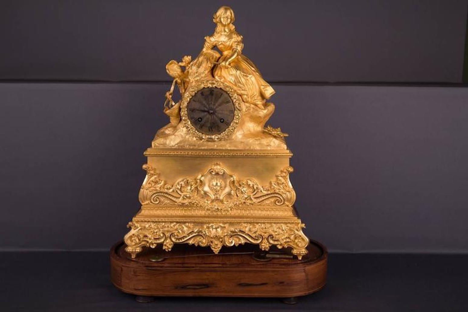 Gran péndulo de bronce. Base y pedestal con profusos adornos florales y figurativos, como rocaille, hojas de acanto, cartelas, guirnaldas, putti y máscaras centrales. Base adicional ovalada de madera sobre cuatro pies. Con llave de ascensor. Ocho