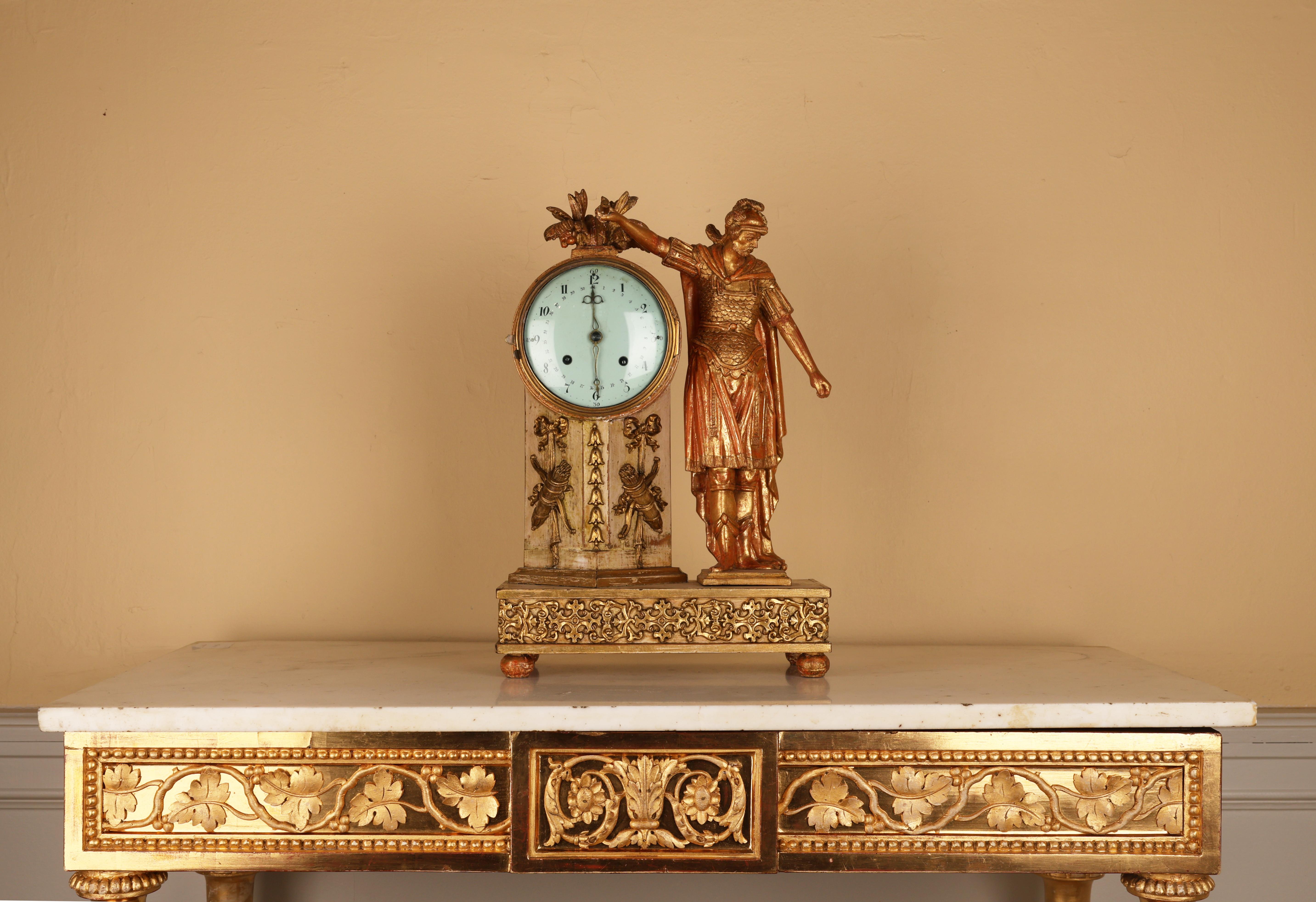An Empire mantel clock in gilded wood, made in Austria or Germany 1820-1830. It features a warrior dressed in classical armor with his arm resting atop the clock. The clock pillar is adorned with bows and arrows and the base is decorated with