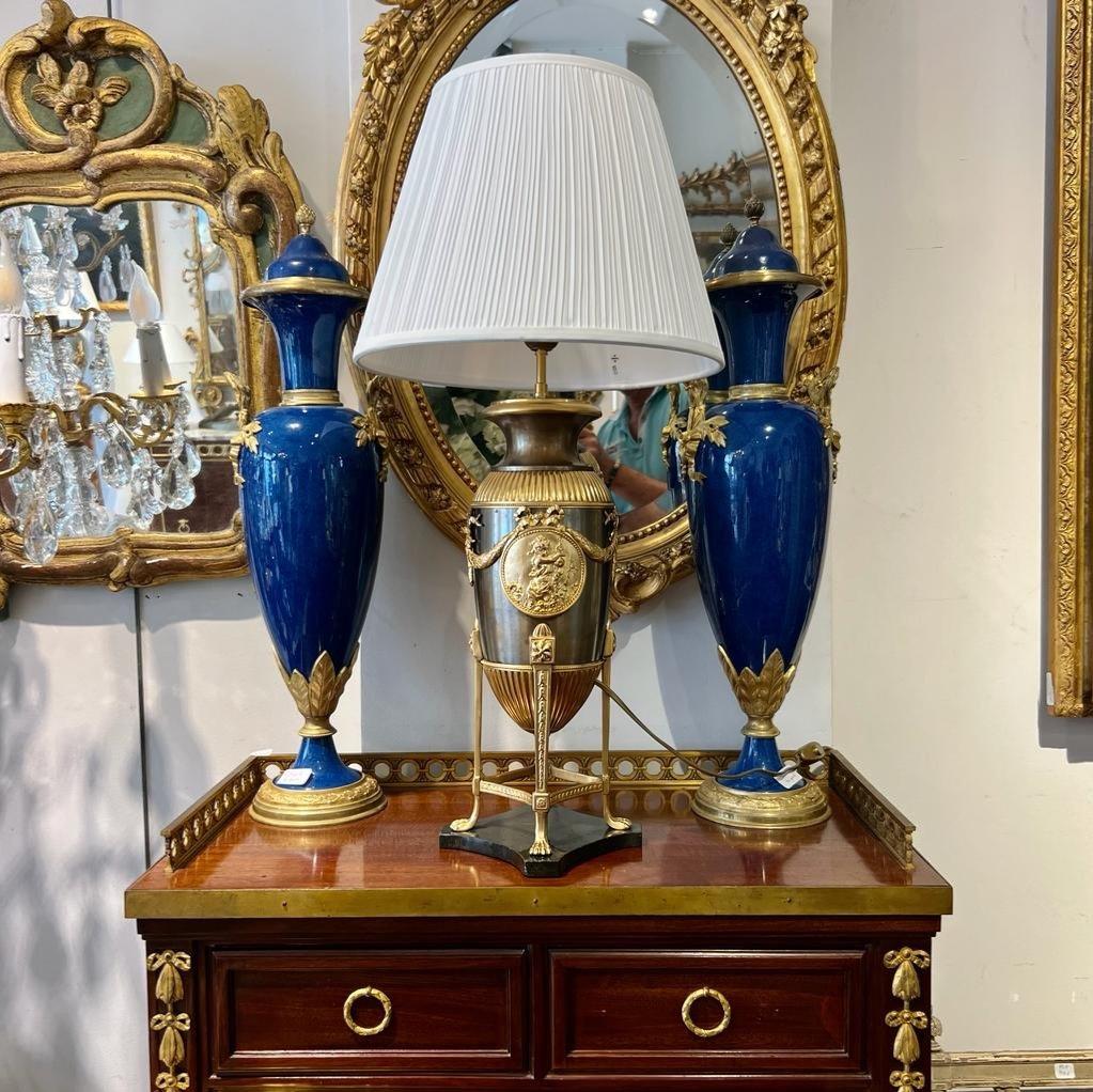 A large four-legged lamp in the style of the First French Empire with a gilded bronze and brown patina finish, resting gracefully on a marble base. Adorned with intricate garlands and two medallions featuring a cherubic figure, this lamp boasts an