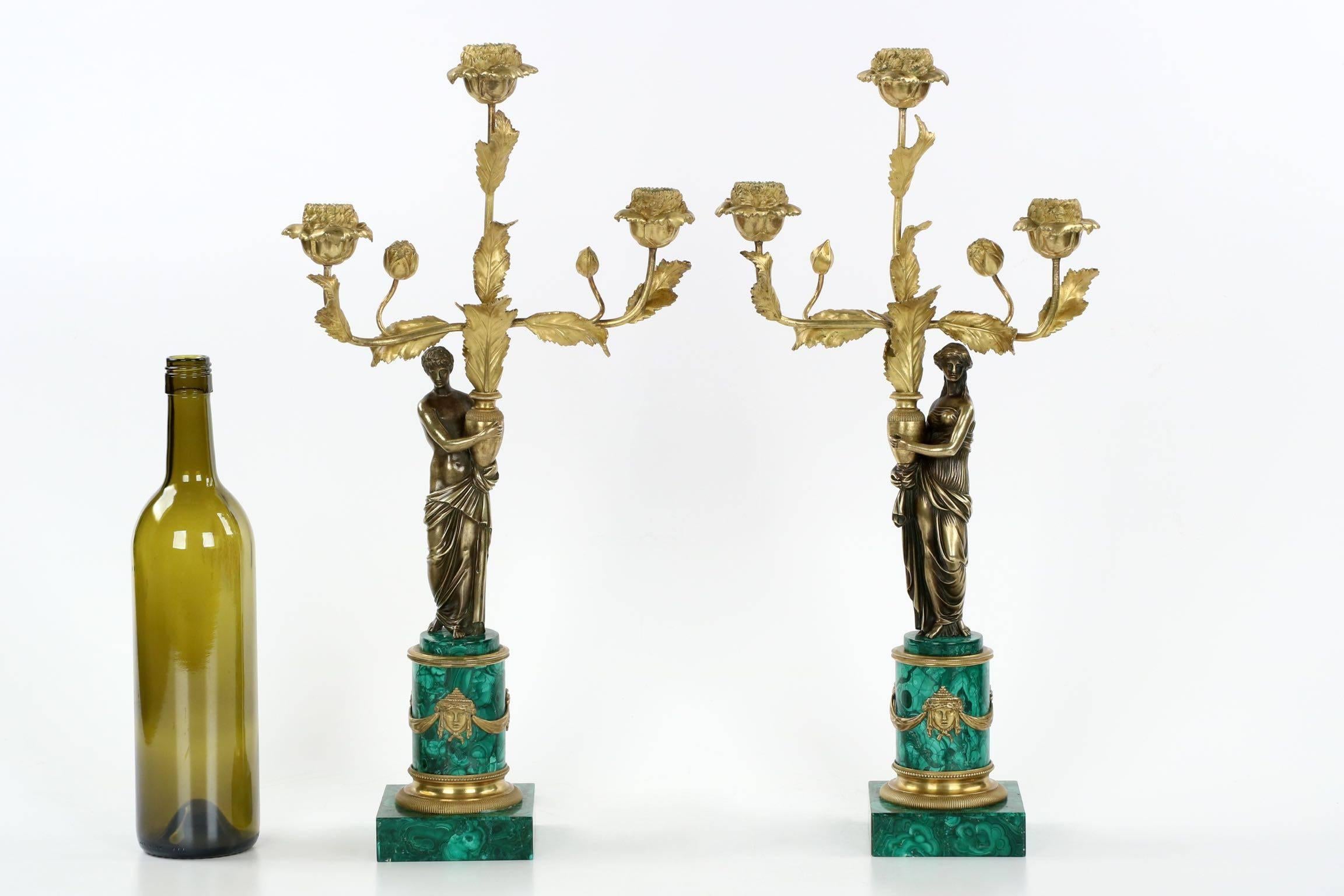 A most attractive pair of late 19th century candelabra in the Empire taste, the male and female figurals raise gilded stems of serrated foliage and budding flowers, the tips culminating in three fully developed flowers holding candle wells. The pair
