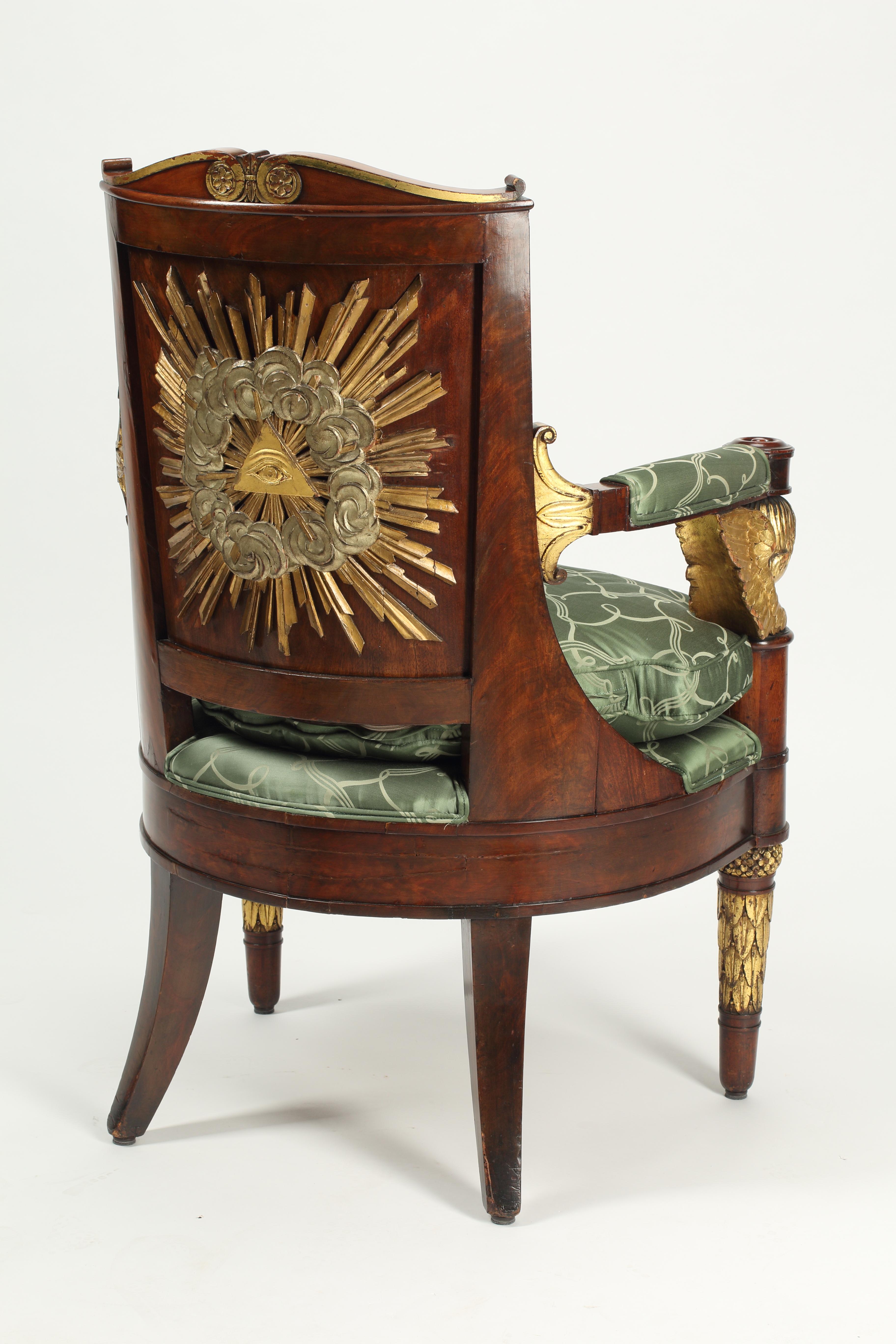 A stately 19th Century Empire Style armchair crafted from Mahogany with detailed gilt carvings and a neoclassical print green silk upholstery. Featuring a gilt sunburst and Eye of God motif on the back of the chair and impressive gilt cherub arm