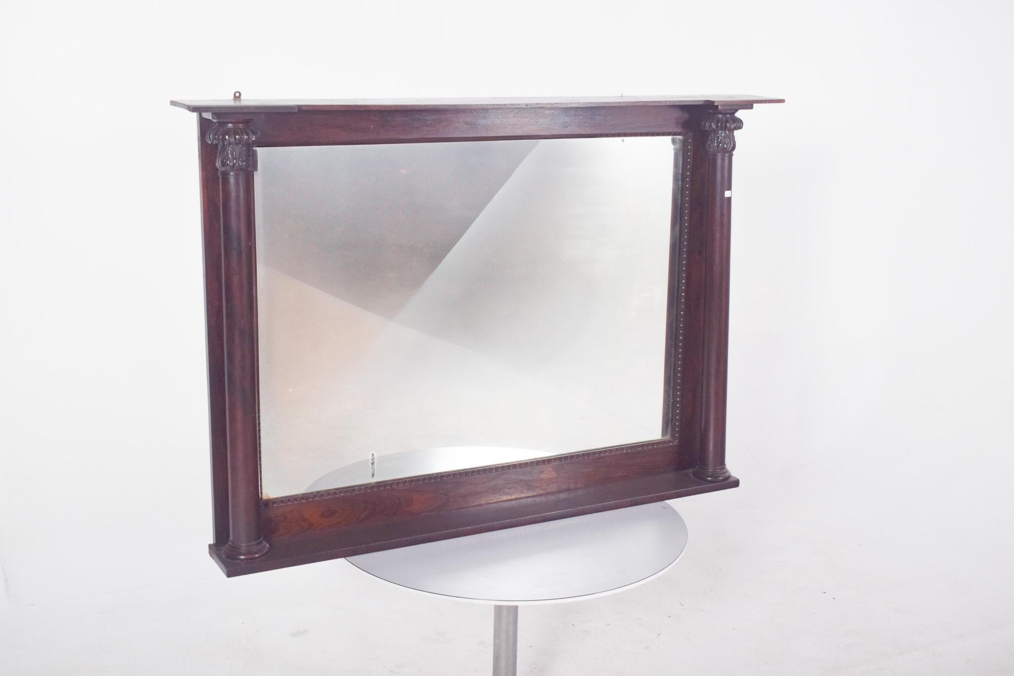 Espejo rectangular estilo Imperio en madera de palisandro. De madera tallada, con un armazón robusto y una presencia impactante, la estructura presenta dos columnas verticales estriadas en los laterales, con capiteles decorados, que recuerdan a la