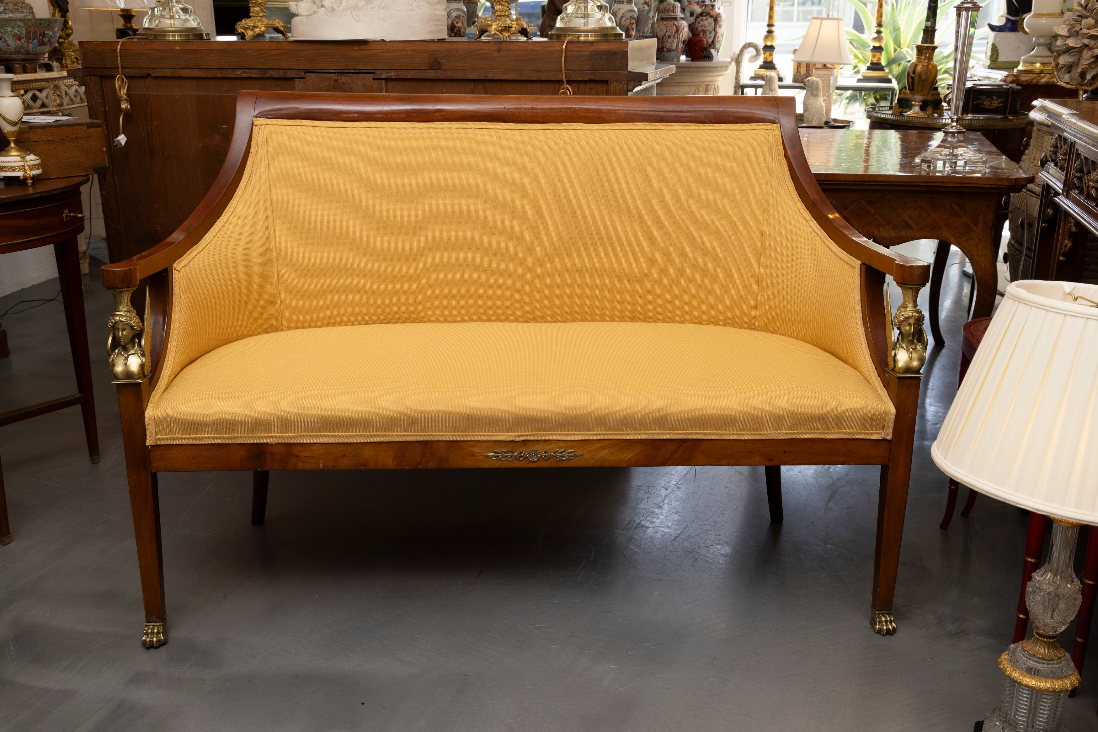 This is a lovely tailored walnut Empire settee.  The top rail is flanked by two graceful sloping arms, joining two brass classical female heads crowing two front straight tapering legs which terminate in brass paw feet.  The settee is fully