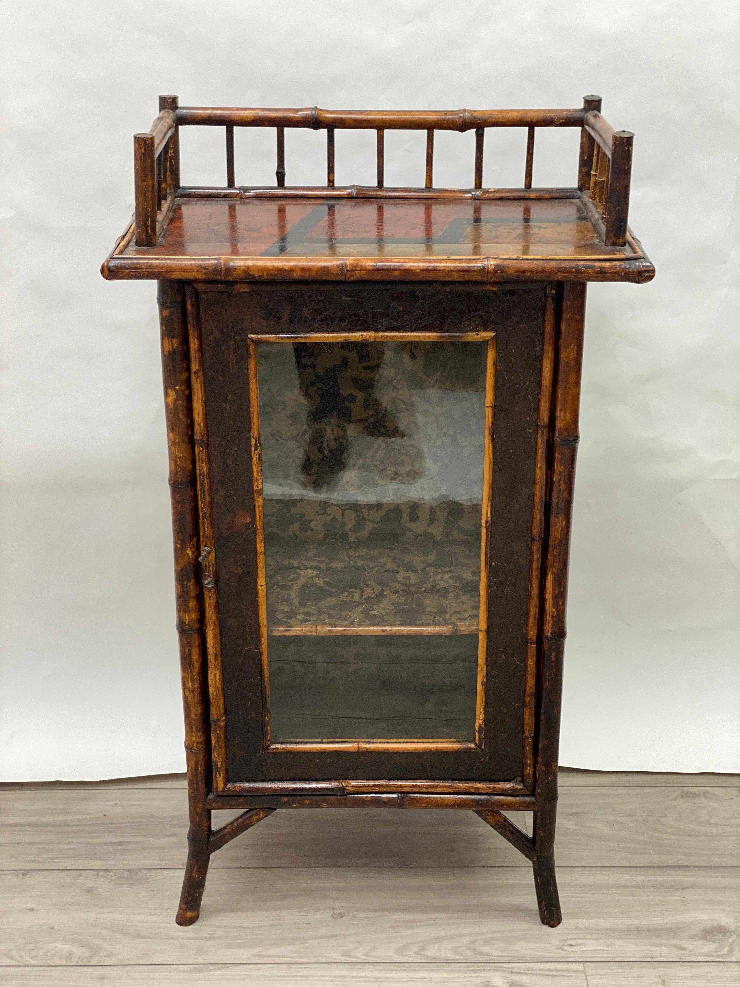 Quaint 19th century English Aesthetic movement bamboo stand. The sides and door are lined with tooled leather. The top surface is lacquered and hand painted, The interior has one fixed shelf and is upholstered. The glass door is orignal to the