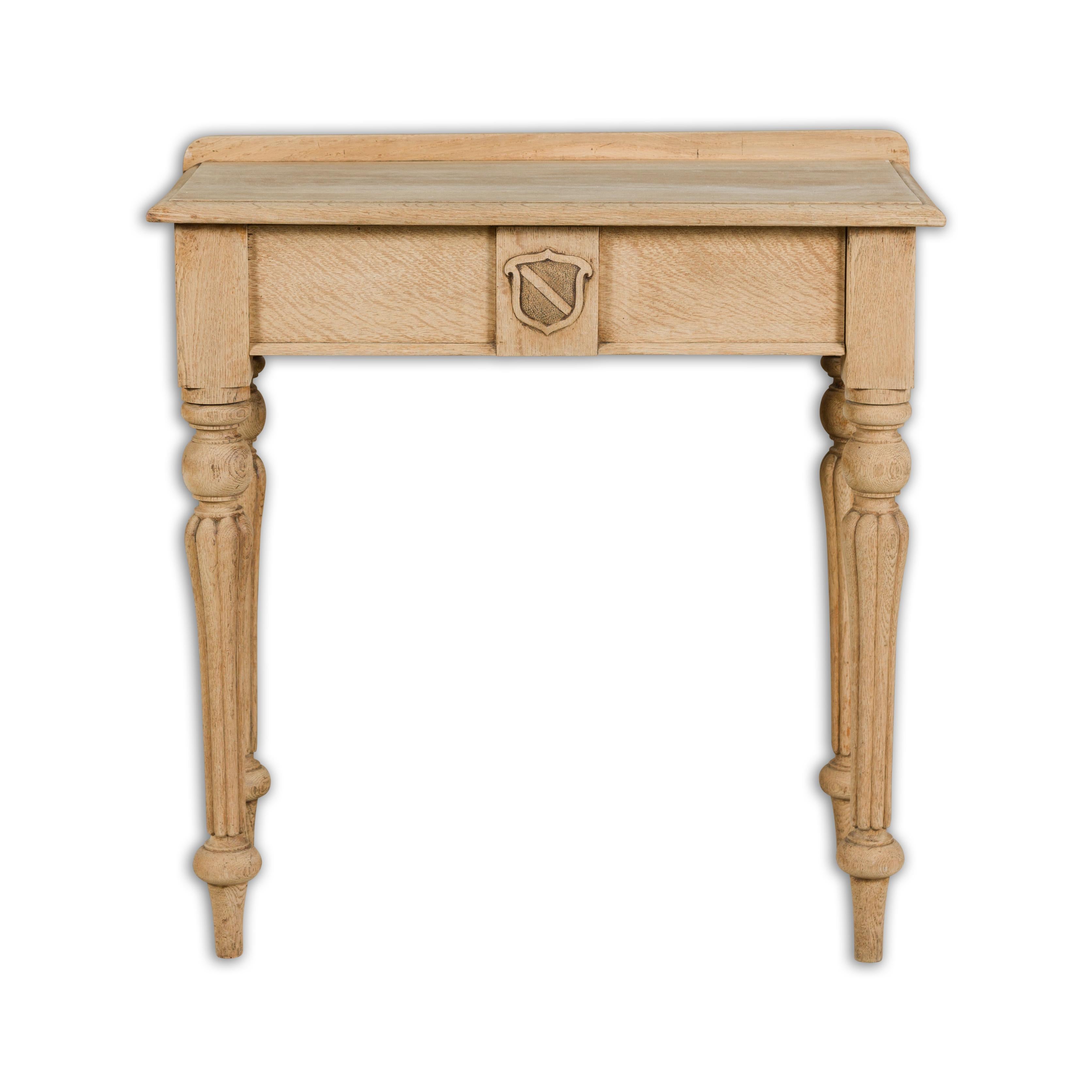 An English bleached oak side table from the 19th century with slightly raised back, single drawer carved with a shield in low-relief, four turned legs adorned with gadroon effects. Introducing a 19th-century English bleached oak side table,