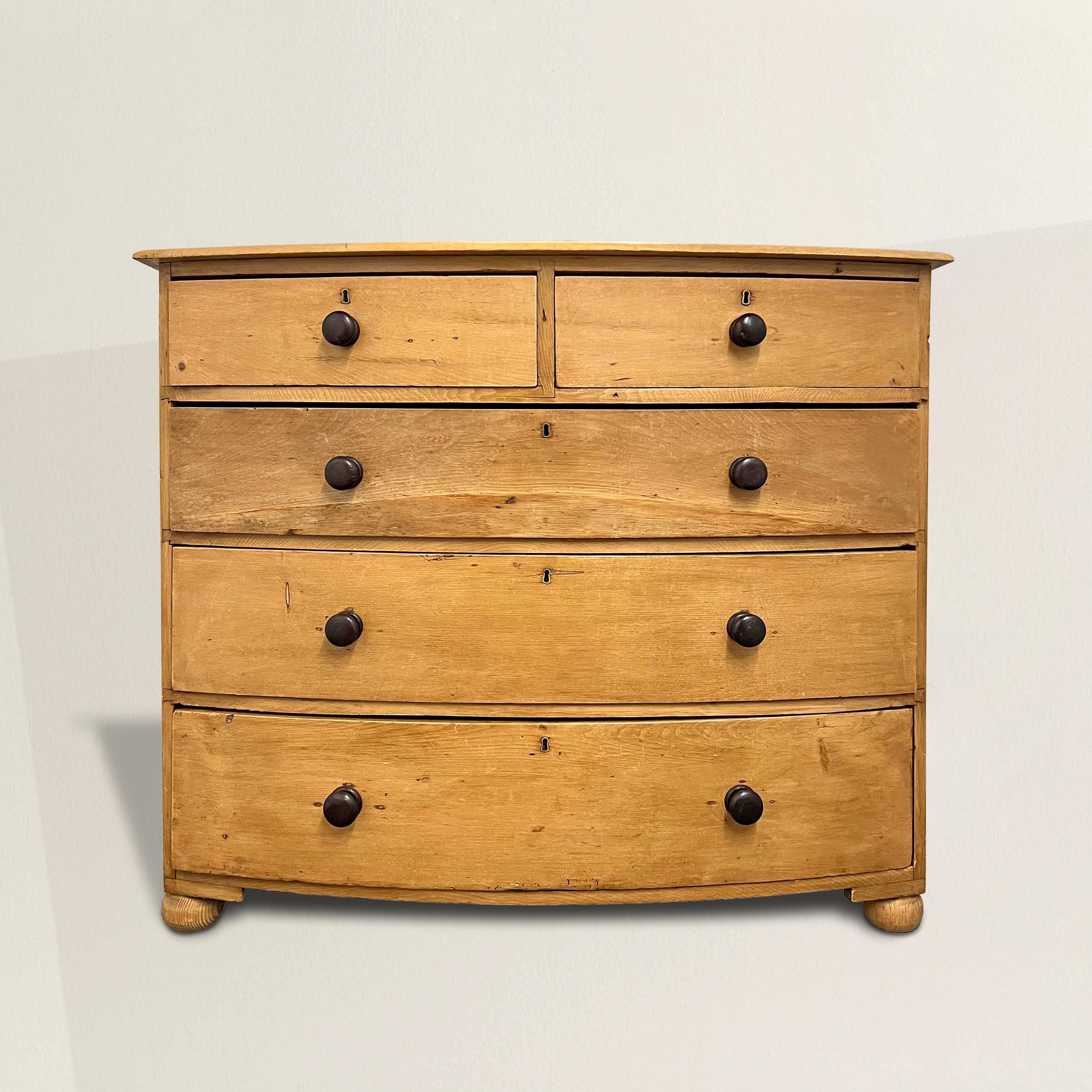 This 19th century English scrub pine bow-front chest of drawers is a remarkable example of English country furniture. With a charming two over three configuration, it offers practical storage and a distinctive aesthetic. The dark stained maple knobs