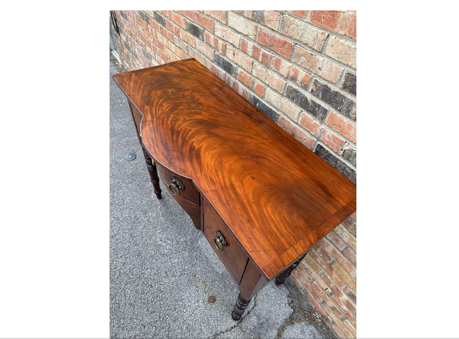 This is a beautiful English brandy board! Dating to the 19th century, it has lovely gloss and patina that accentuates the natural beauty of the wood. There are two deep drawers on either side, with a smaller drawer in the curved center. The legs