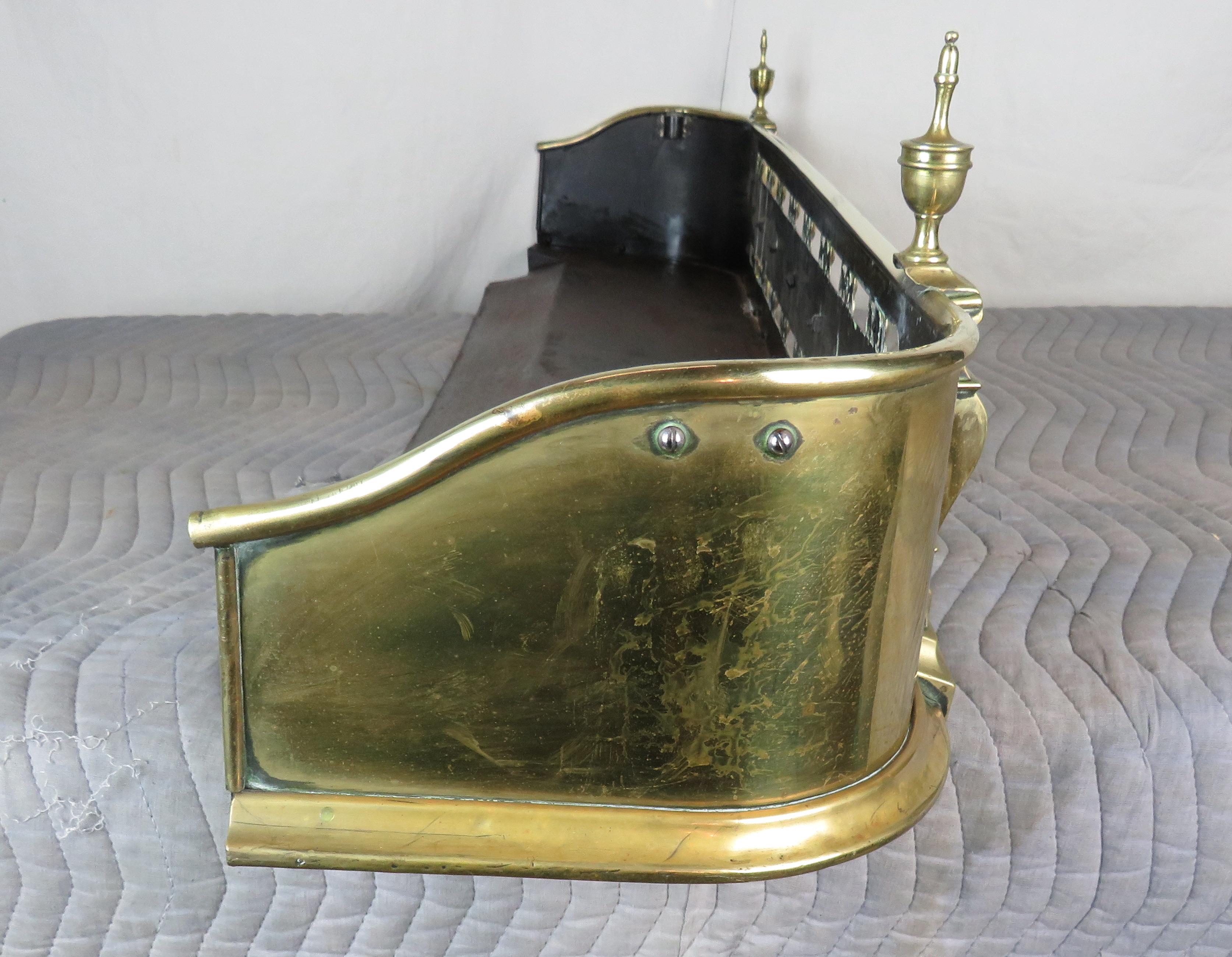 Brass fire fender with urn finials atop shaped molding on either side of pierced front.