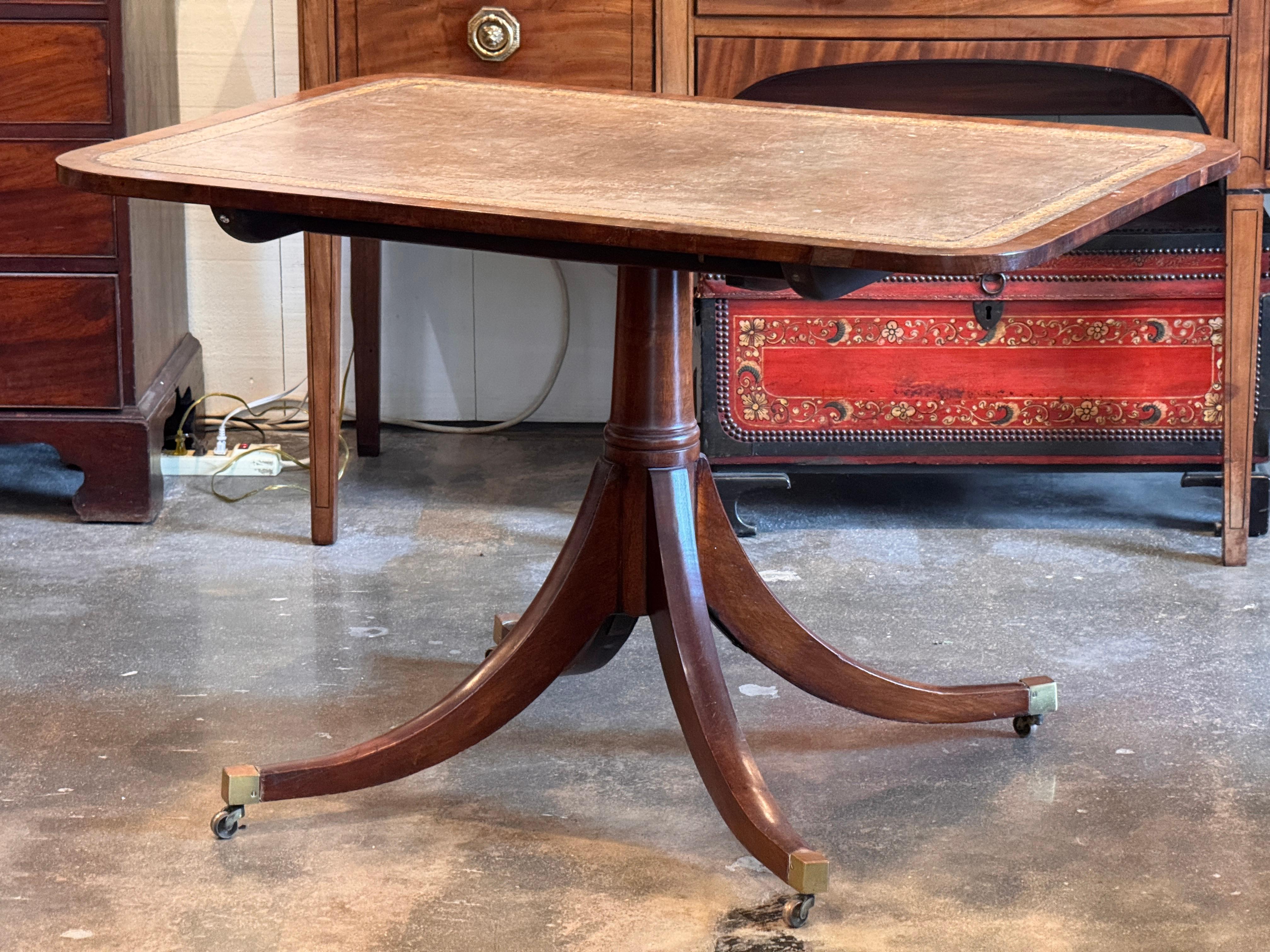 Rectangular tilt-top table with tooled leather surface over a reeded mahogany edge, supported on a turned pedestal base with four splayed legs ending in brass caps and casters. Retains its original brass tilt mechanism and shows warm patina