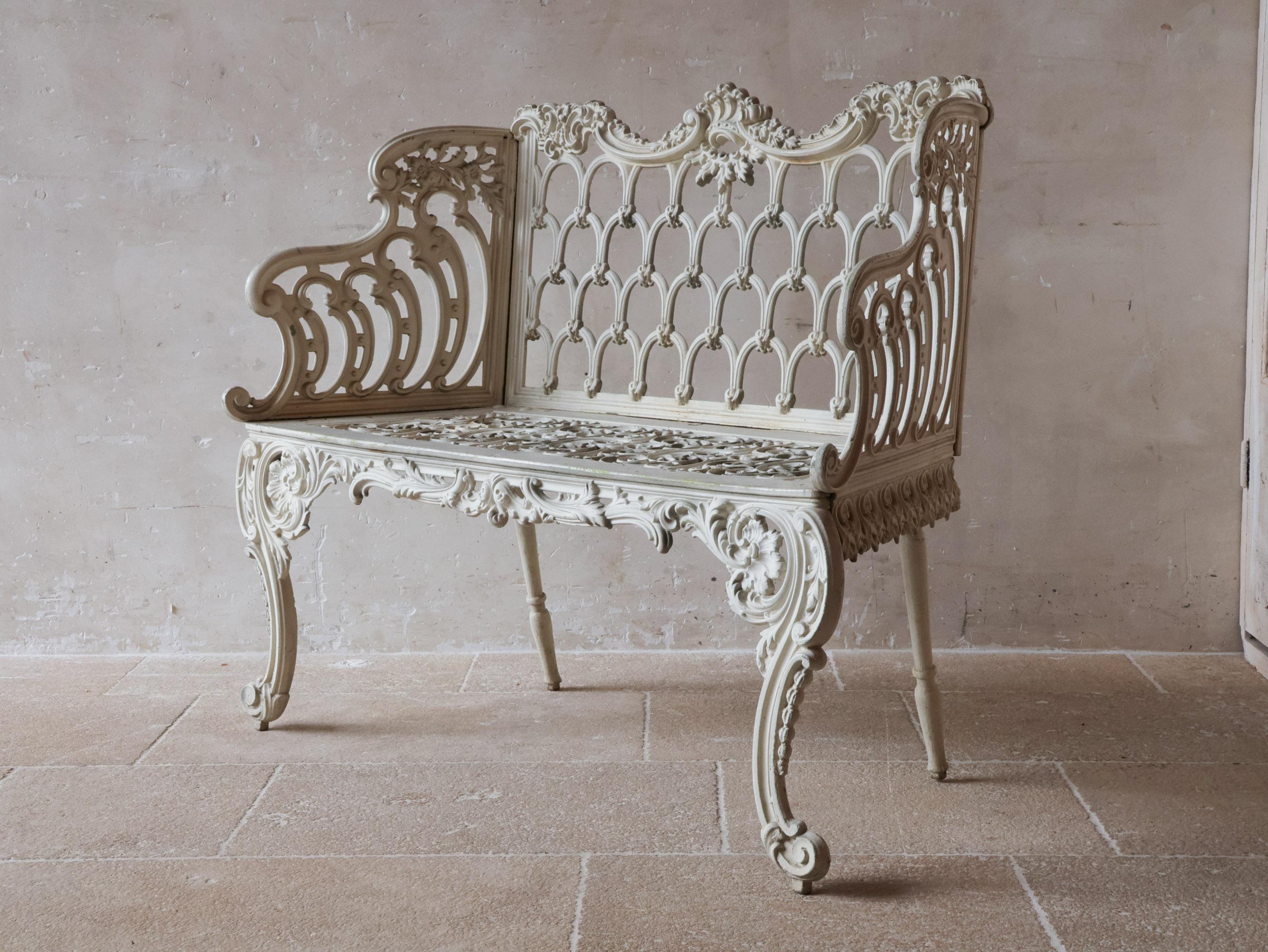 Auténtico y compacto banco de jardín inglés (love seat) de hierro fundido pesado con un diseño romántico y ornamentado. Este antiguo mueble de exterior se caracteriza por detallados motivos de flora y fauna en el respaldo y el asiento. El estilo se