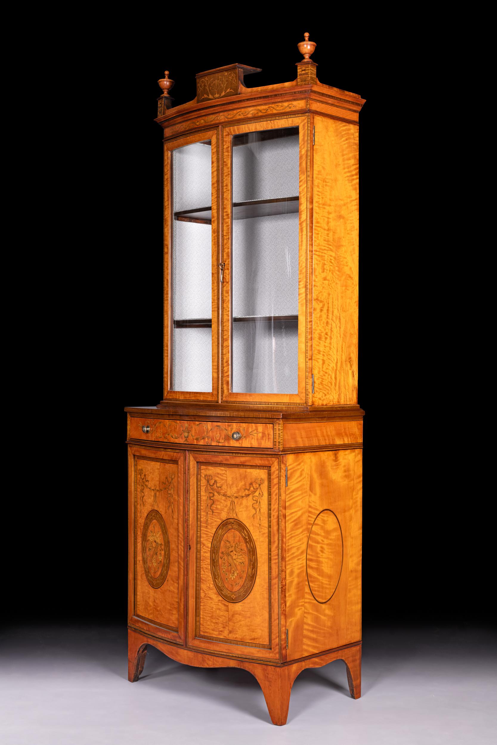 A very fine Edwardian Satinwood bow fronted display cabinet attributed to Edwards & Roberts of London, the upper section with a central pediment flanked by two classical urns above twin glazed bow fronted doors which open to reveal a shelved