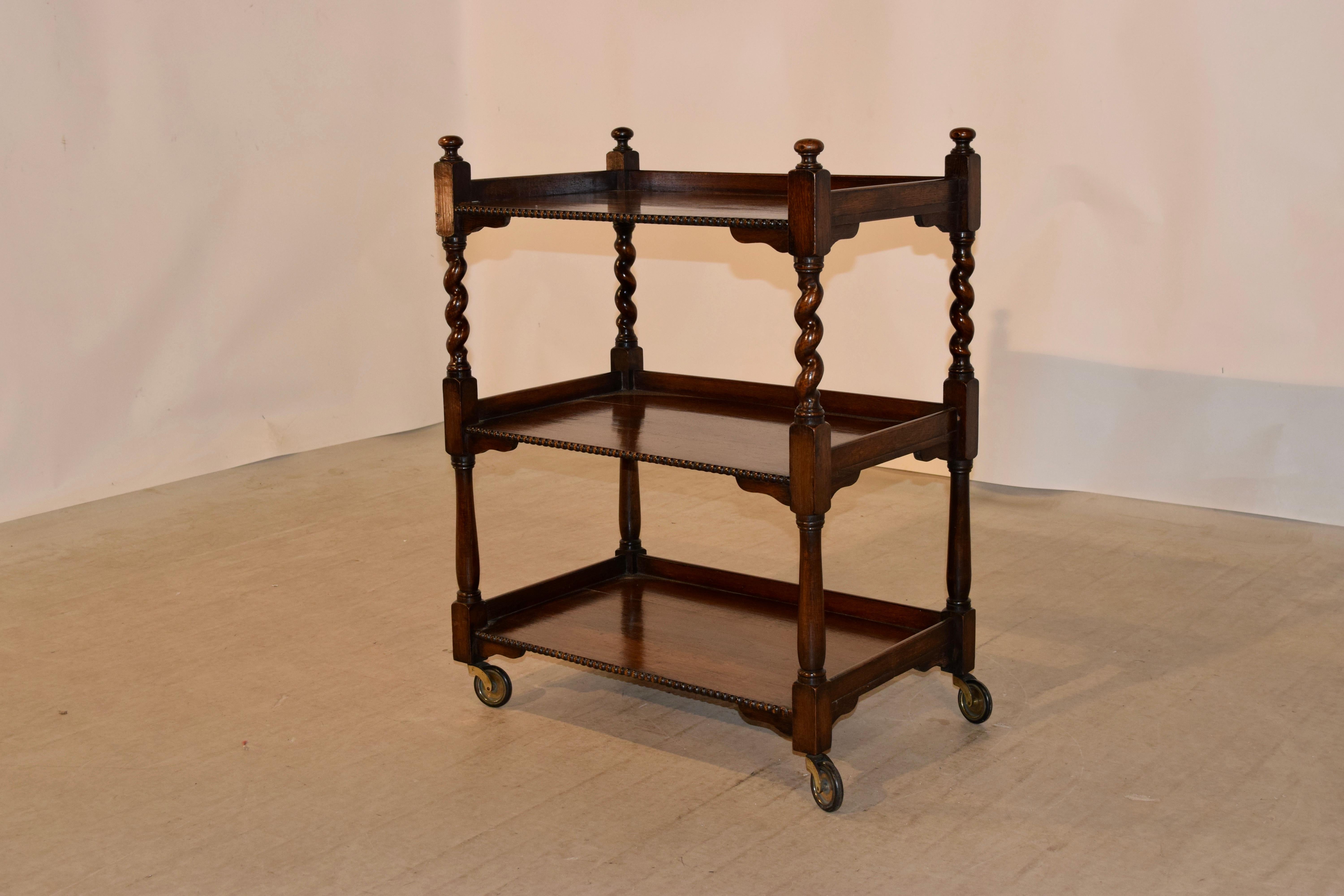 19th century English oak drinks cart with three shelves. The top is decorated with finials, following down to three shelves, all with beaded front edges and galleries. They are separated by hand turned barley twist shelf supports on the top and hand