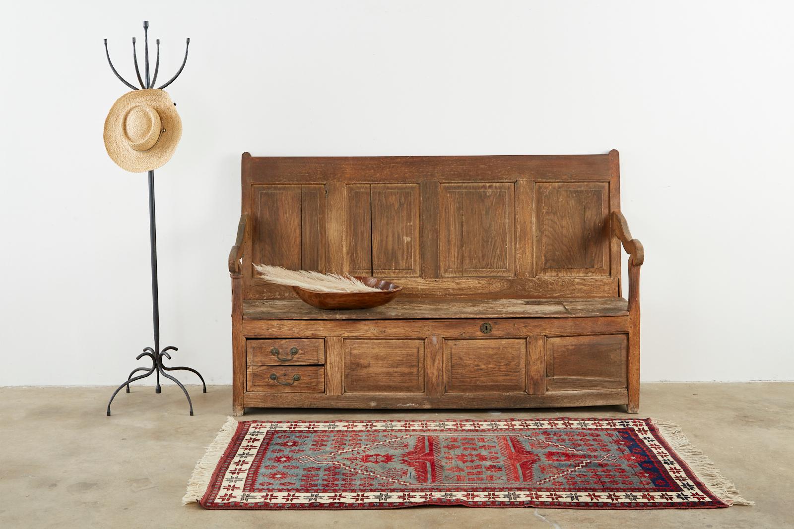 Rustic 19th century English Georgian box settle or hall bench. Crafted from oak featuring a paneled back. The large storage box has two small storage drawers on the front left with iron pulls. The seat has a hinged lid that opens to the large