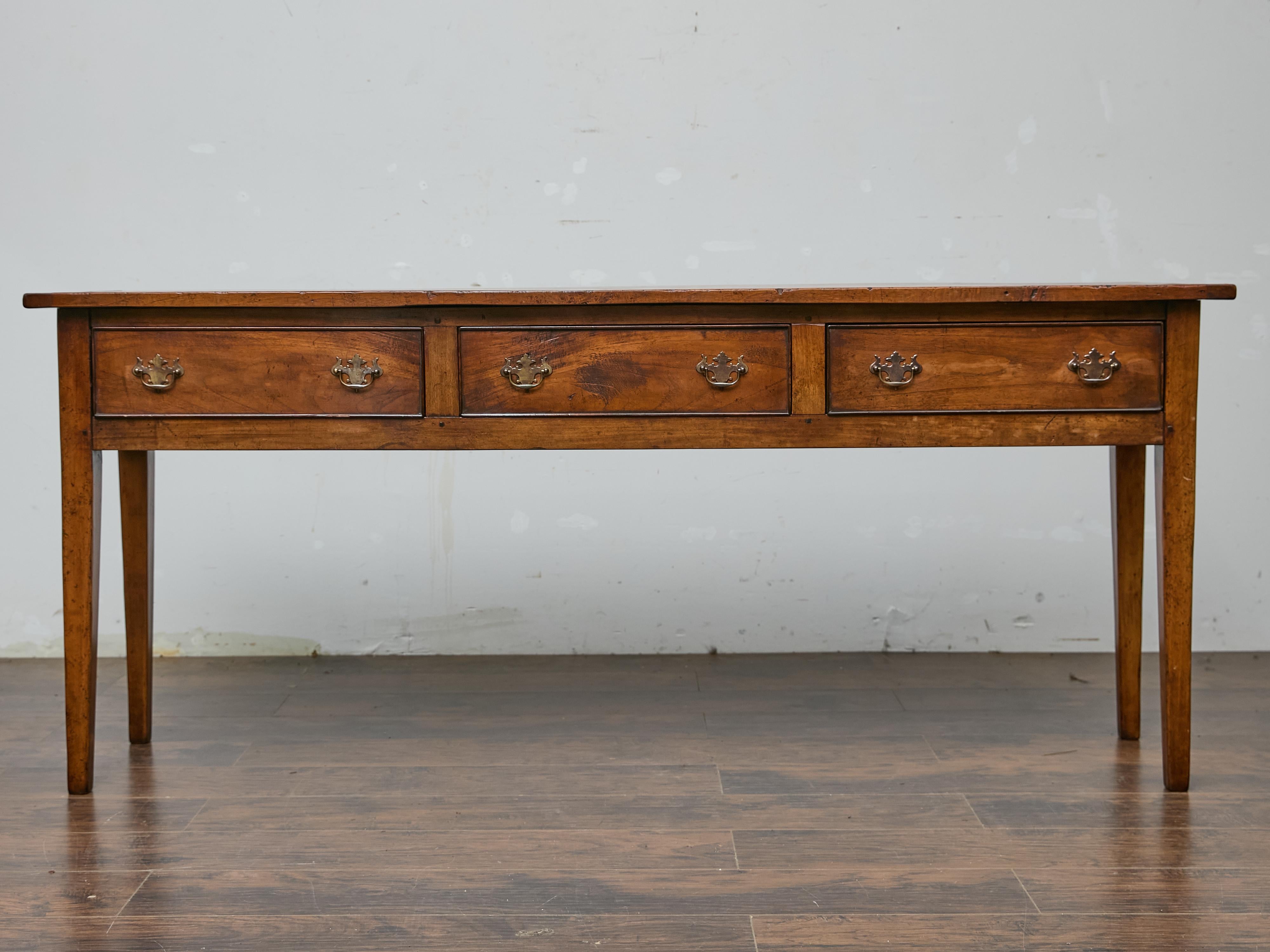 This English Georgian server table in pine, dating to the 19th century, is an understated yet elegant piece that brings both function and character to a room. Crafted with a long and narrow form, it features three spacious drawers adorned with brass