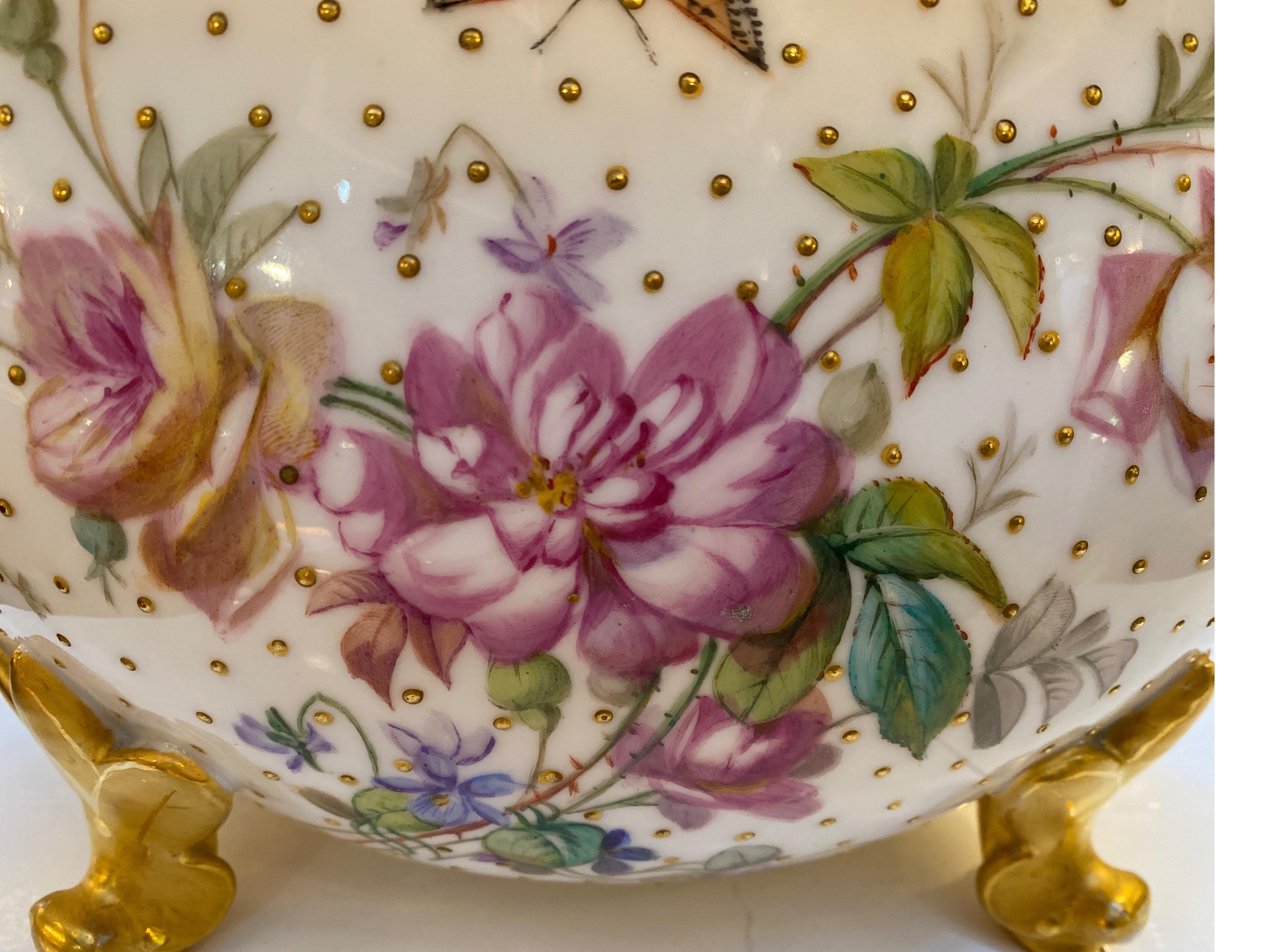 Elegant vase sur pied de forme ovale en porcelaine peint à la main avec décoration florale. La bordure dorée perlée est ornée d'une peinture à la main florale sur toute sa surface, avec des perles en relief sur le corps. Quelques insectes et