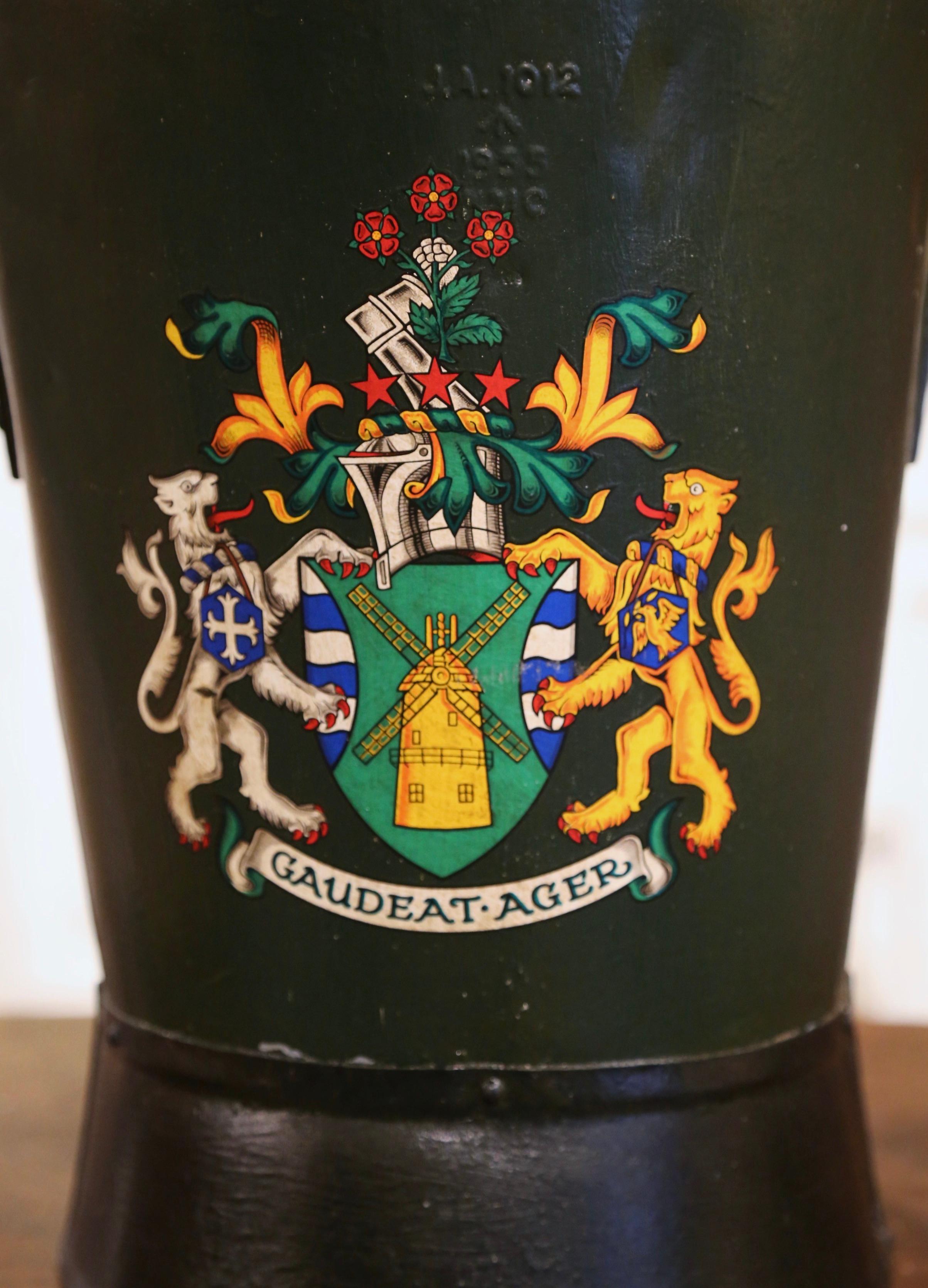 Place this large, colorful antique stand at your front door or in your mudroom to collect umbrellas, canes or other accessories. Created in England, circa 1890, the iron bucket with top and side handles is decorated with a coat of arms with armor,