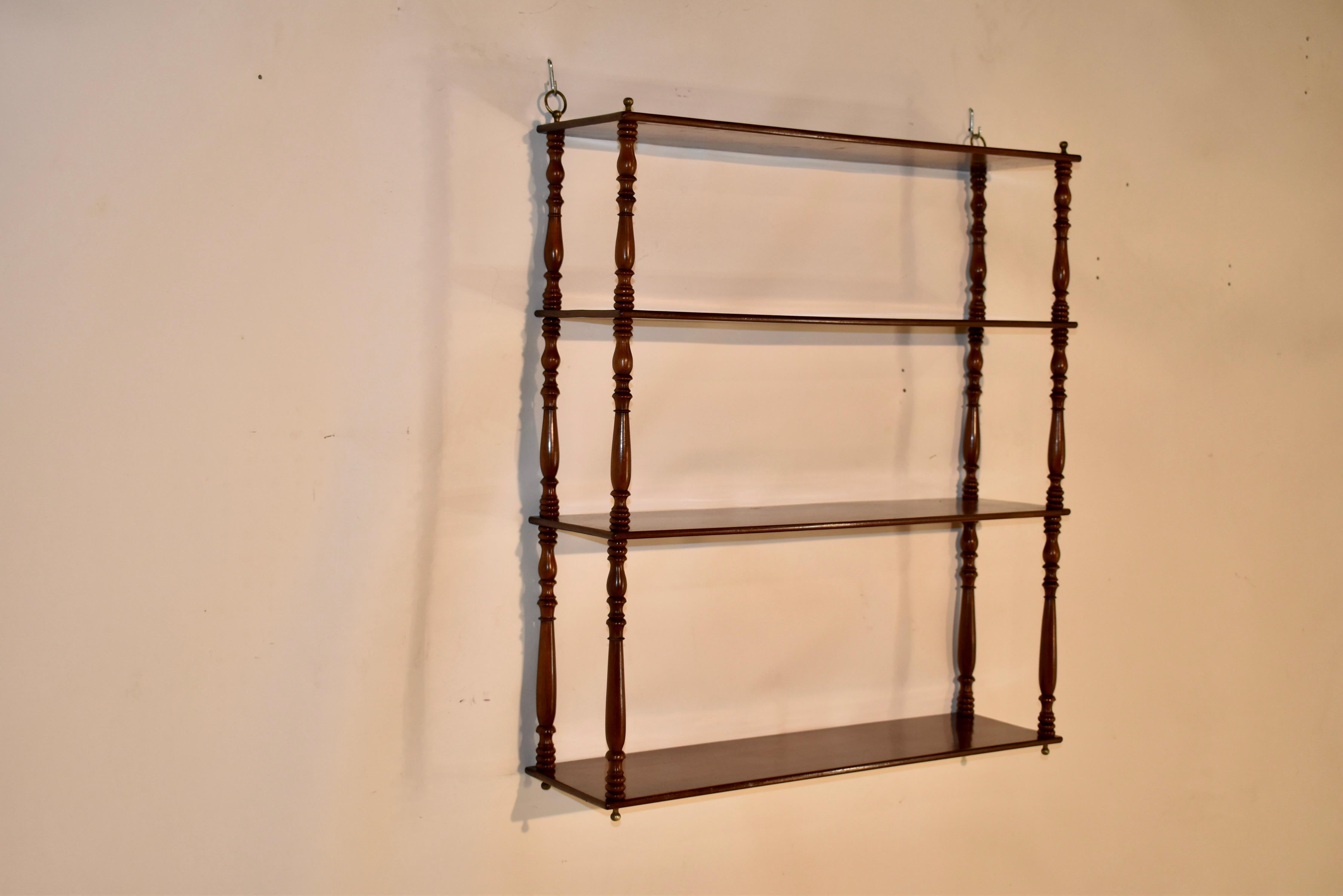 19th century mahogany hanging wall shelf from England. The hanging shelf has four shelves, all single mahogany boards, separated by hand turned shelf supports and decorated with brass finials on the top and bottom. Original hanging rings on the top