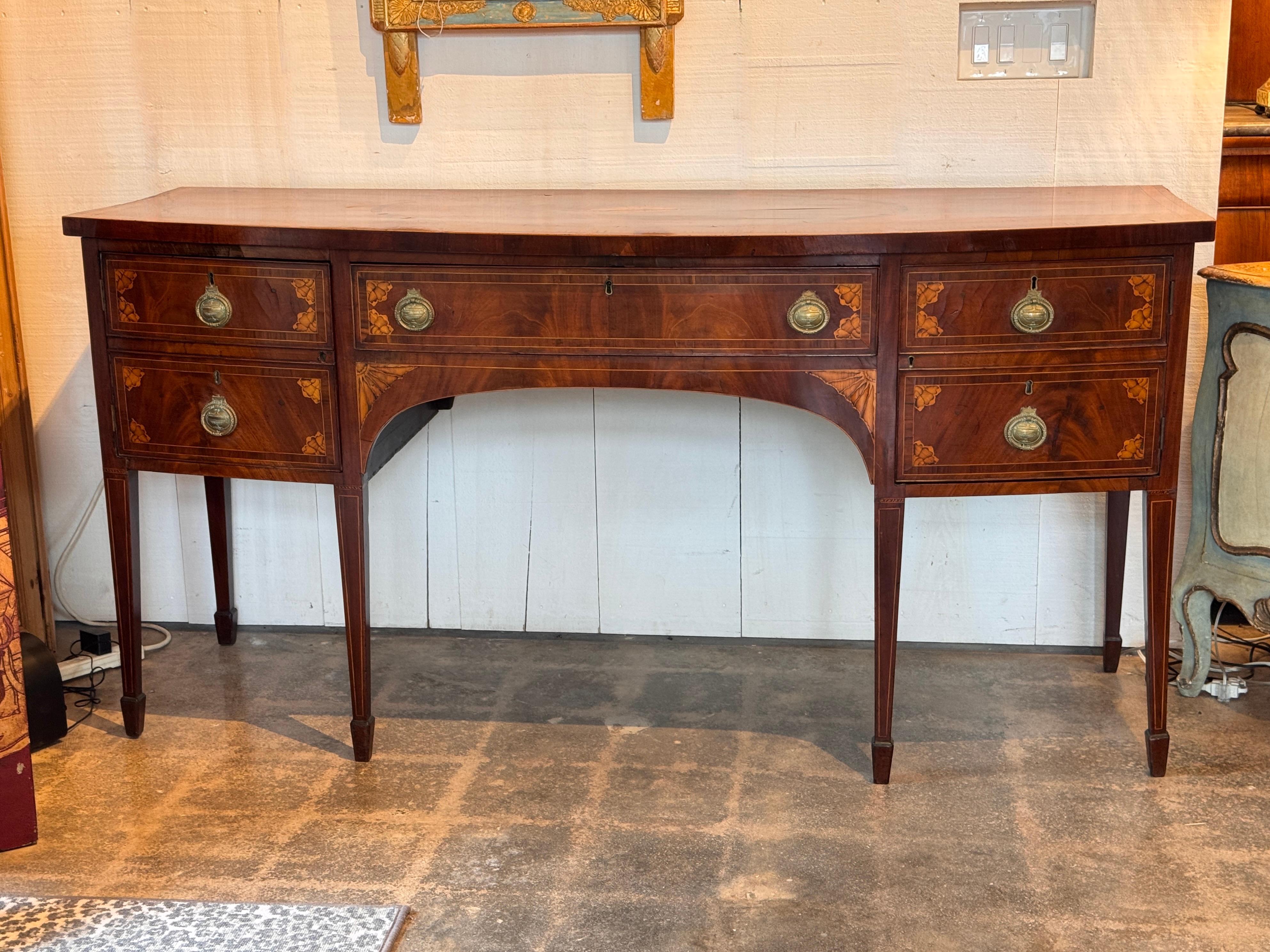 Credenza inglese Hepplewhite con frontale a serpentina e raffinati intarsi. Il piano è decorato con un medaglione centrale a ventaglio e un ovale a fasce incrociate delineato da un'impiallacciatura a contrasto. I frontali dei cassetti e le gambe