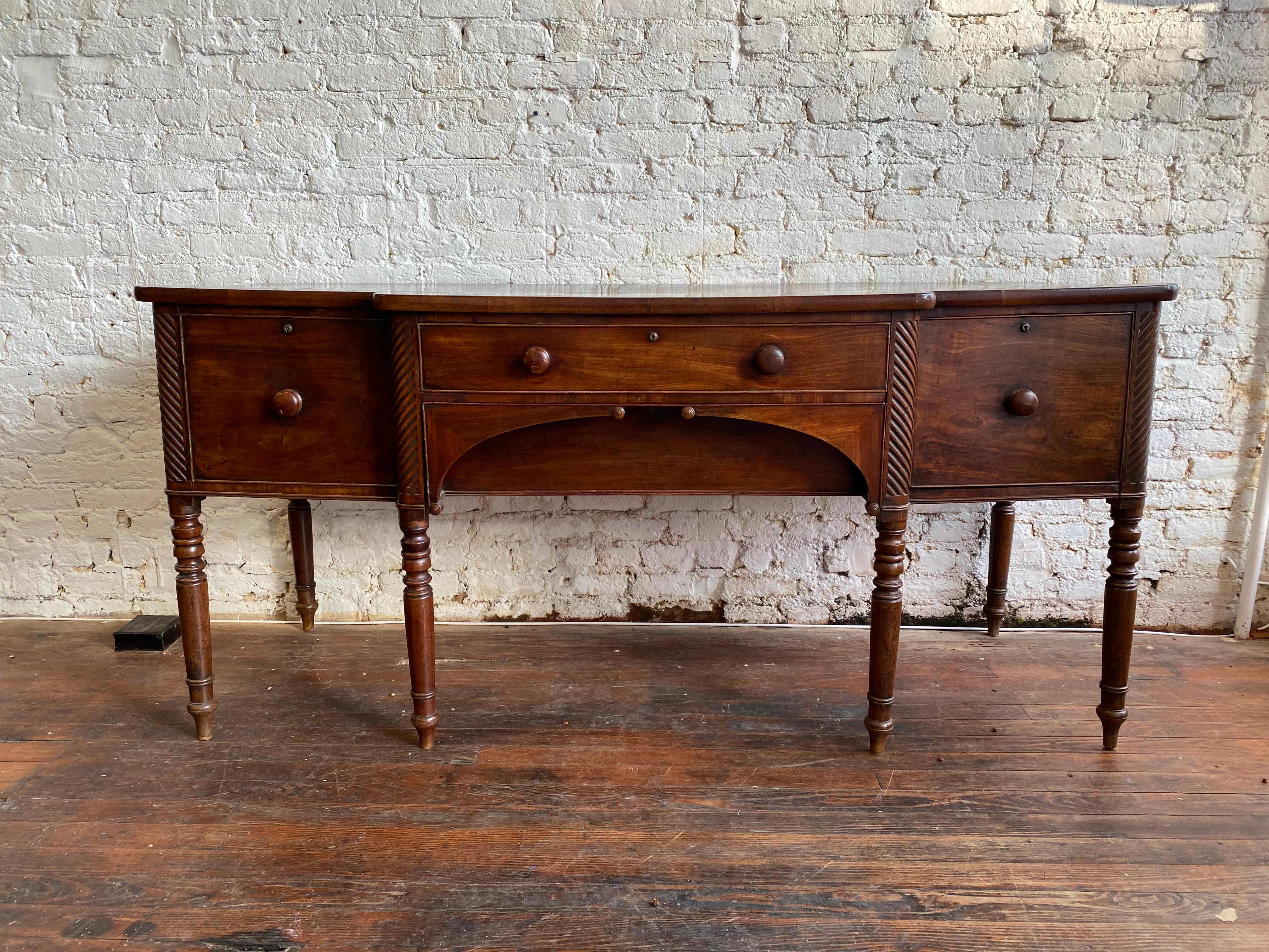 Great color and patina on this 19th century English mahogany breakfront form sideboard with 2 drawers flanked by 2 cabinet doors. The bottom drawer is recessed behind two symmetrically arched handles terminating into hand carved finials.