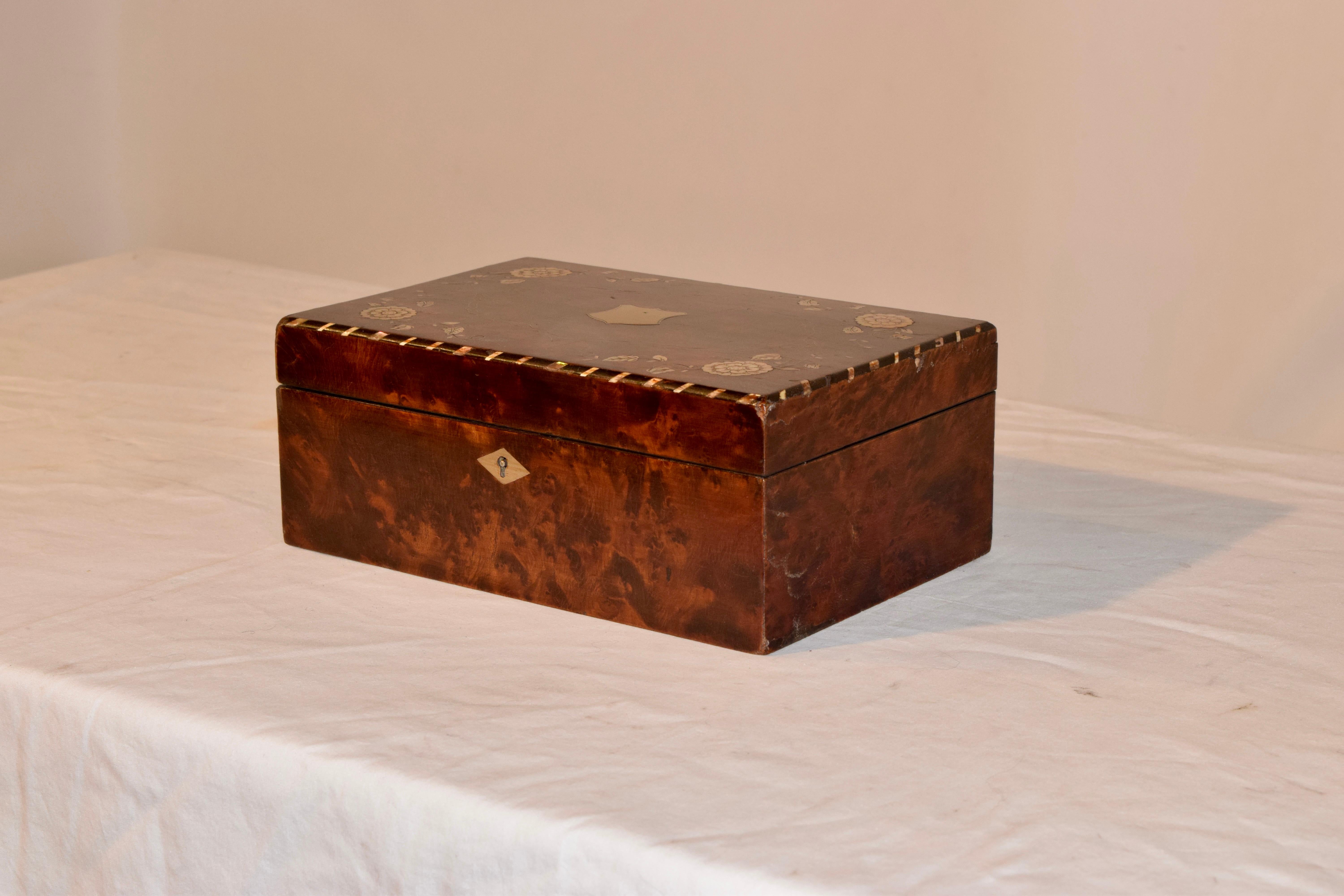 19th century English dresser box made from plum pudding mahogany. The box is intricately inlaid with mother-of-pearl and bone accents. There is a brass shield imbedded in the lid, surrounded by lovely florals and vines. The top opens to reveal