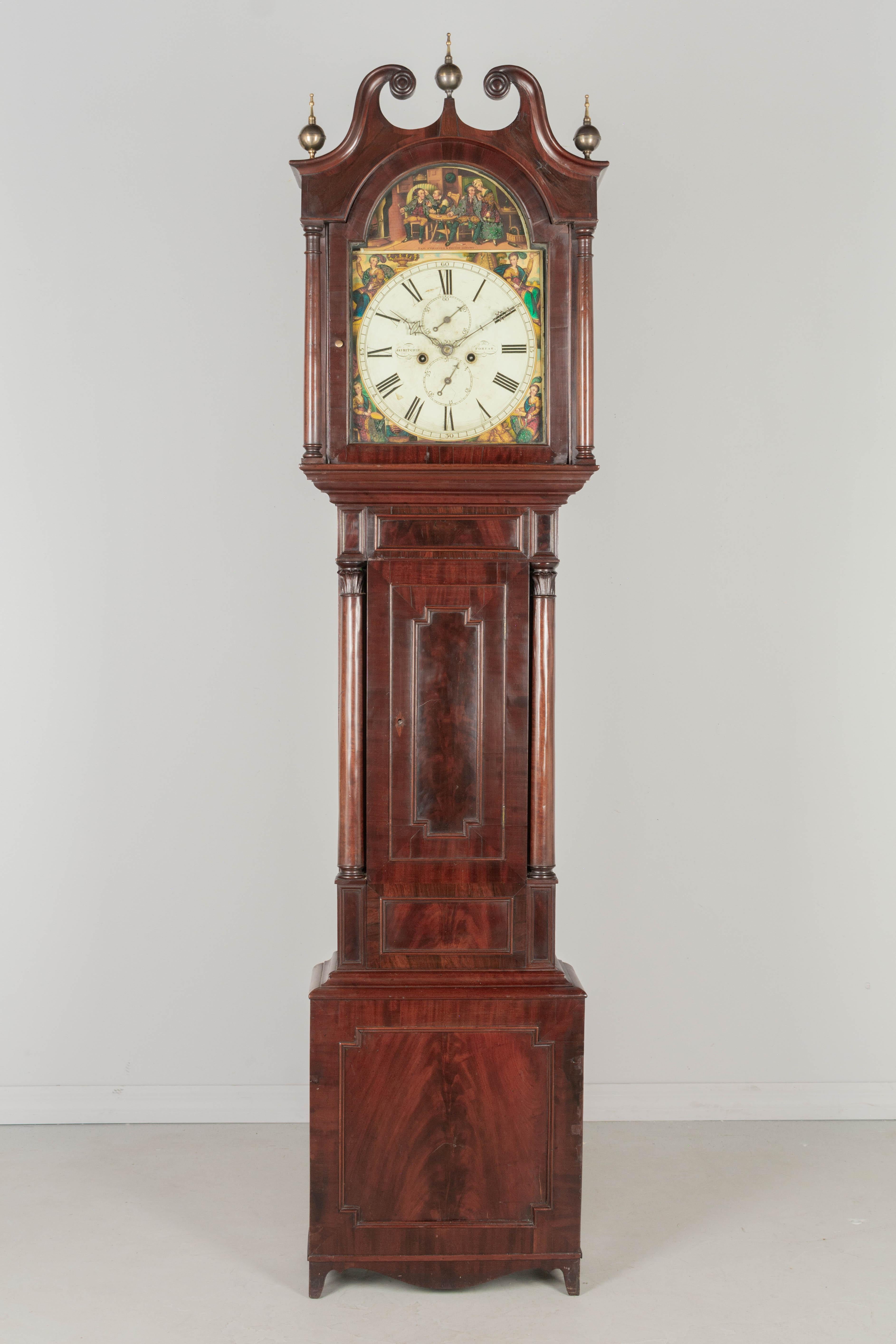 A 19th century English tall case clock crafted of beautiful figured and crotch, bookmatched mahogany with turned columns flanking dial case and door. Decorative brass ball finials on carved split-pediment top. Colorful painted surround illustrates a