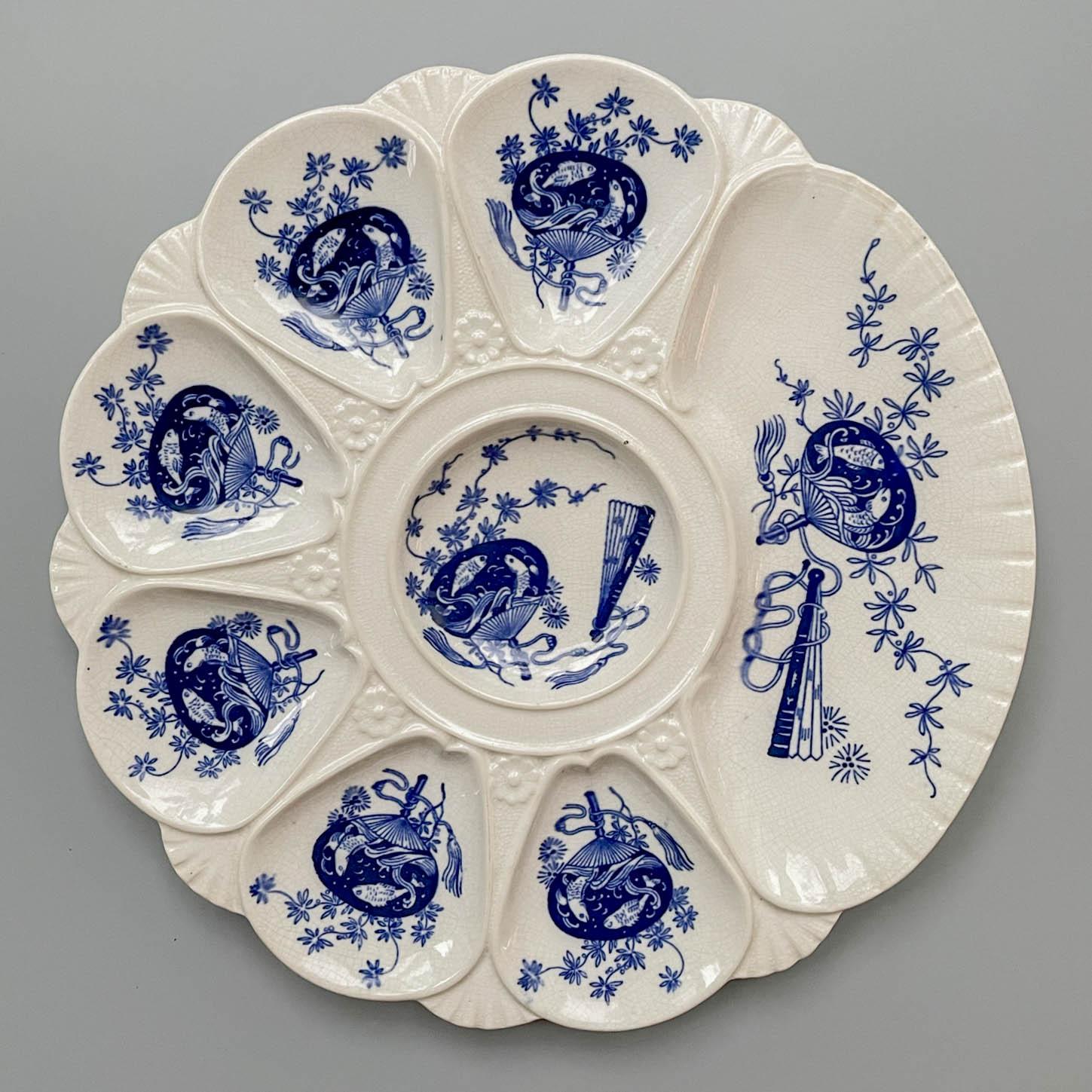 A Victorian Minton flow blue porcelain oyster plate, with six oyster wells, a center well and a large well for crackers. Decorated in the Japonisme style, each well has indigo blue flowers and fans with koi fish and is separated by relief of shells