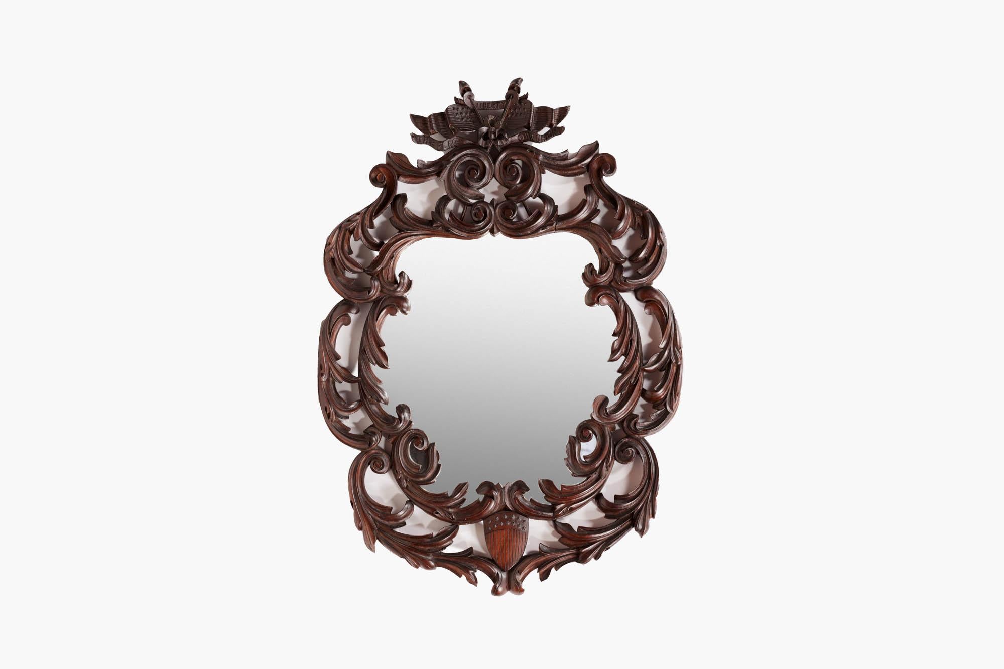 19th century English mirror in the Americanised fashion. The shaped glass plate sits within a heavily carved dark wood frame with foliate scrolling along with a crossed flag emblem at the top and shield detailing at the bottom. The slogan on the top
