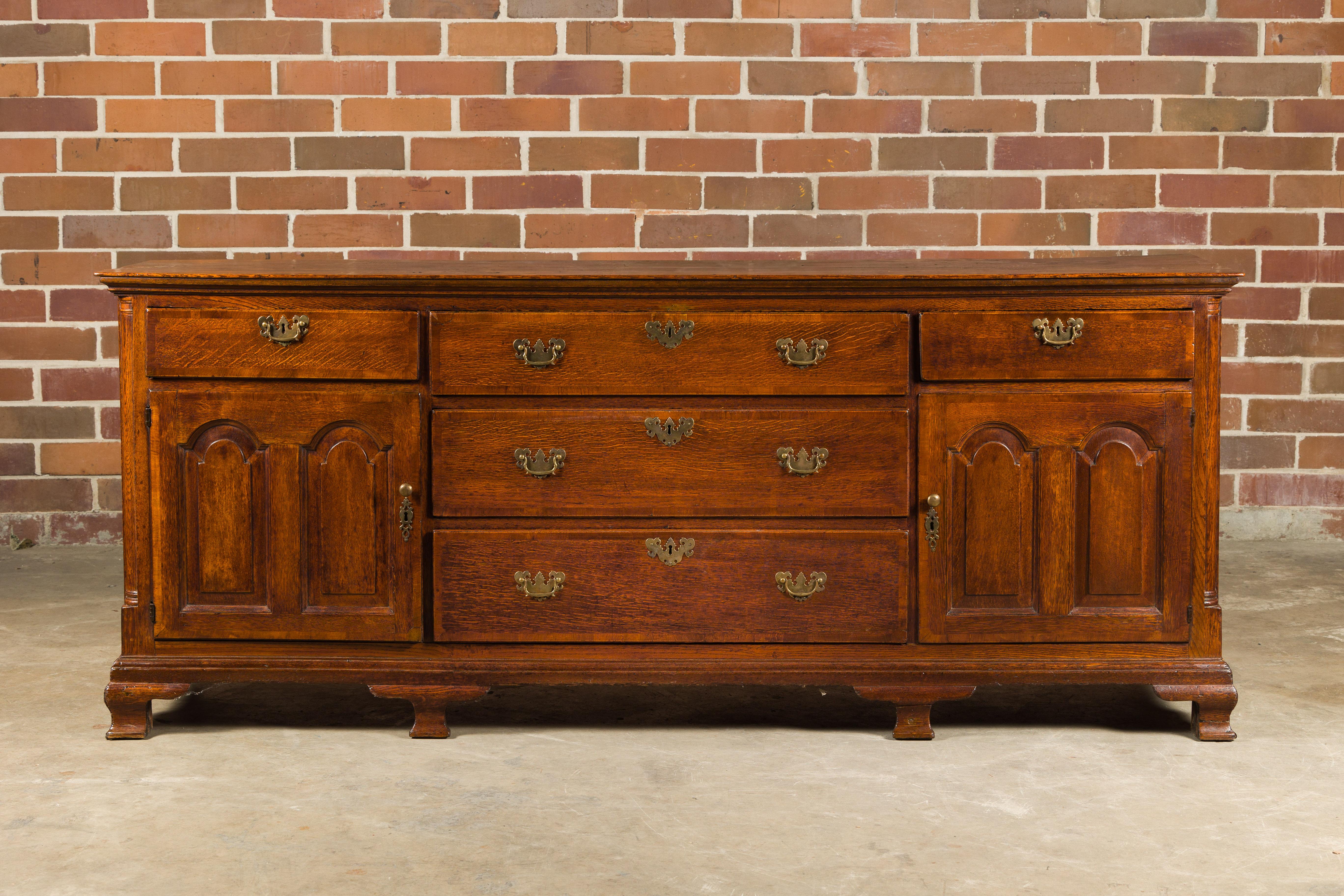 An English oak long buffet from the 19th century with five drawers, two doors, brass hardware and carved feet. Step back into the splendor of 19th-century England with this distinguished oak long buffet. Radiating undeniable antiquarian charm, its