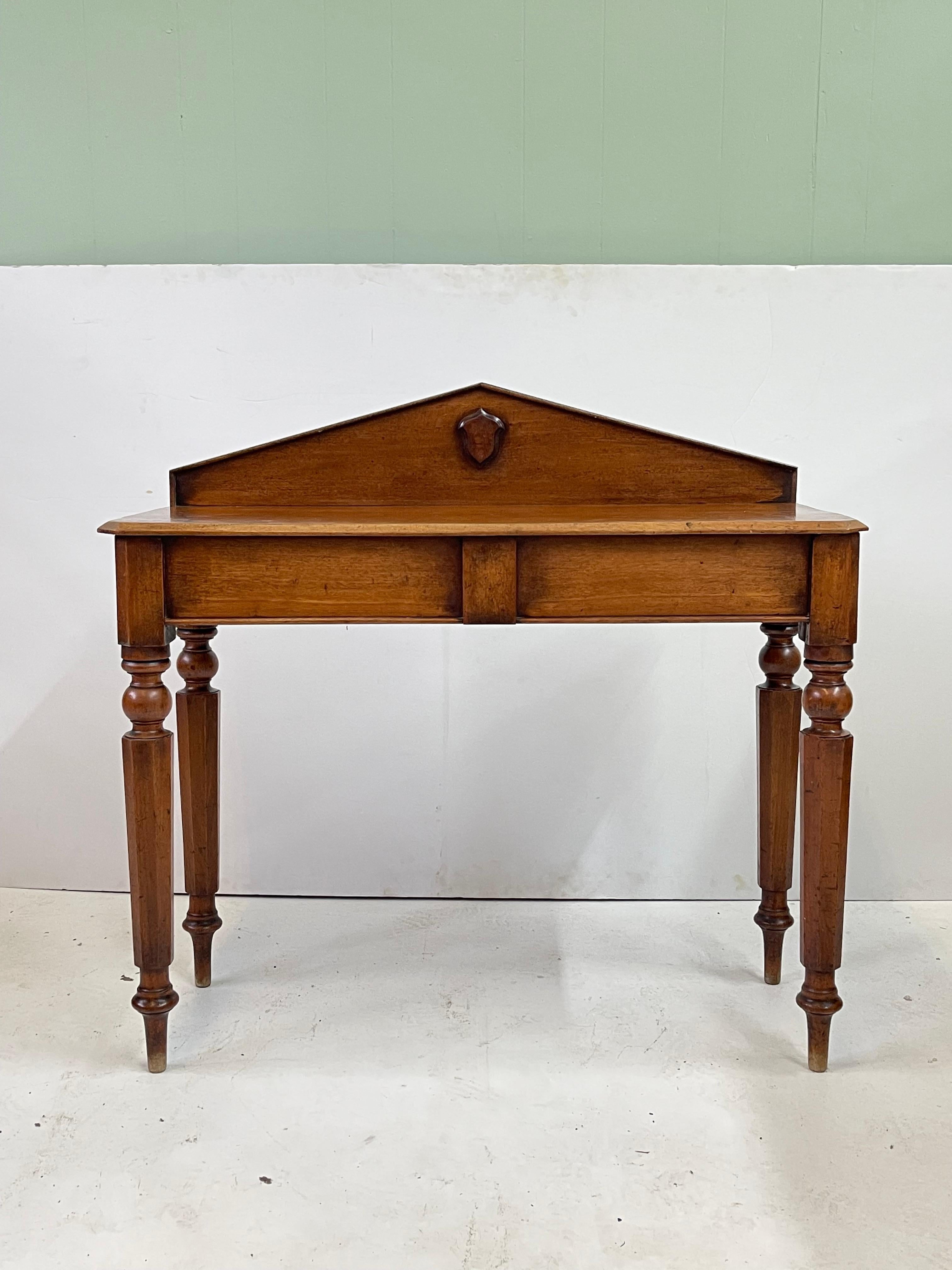 A 19th Century English console table made from oak with a peaked backsplash decorated with a wooden shield. This table features a rectangular top with beveled edges and has a single dovetailed side drawer ingeniously hidden in the apron. The table