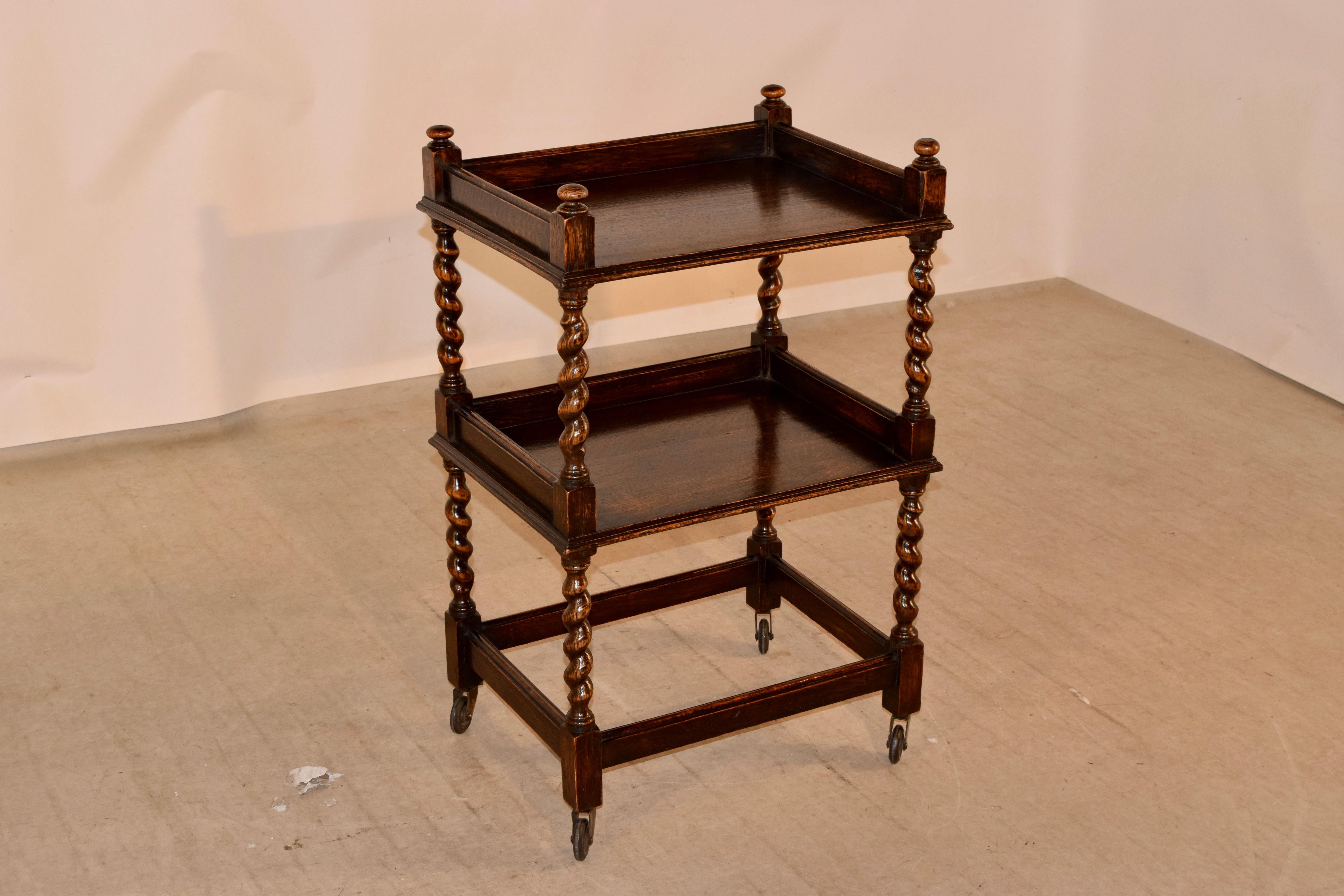 19th century oak drinks cart from England with finials on the top, following down to galleries around the two shelves, separated by hand-turned barley twist shelf supports. The cart is supported on matching hand-turned barley twist legs, joined by