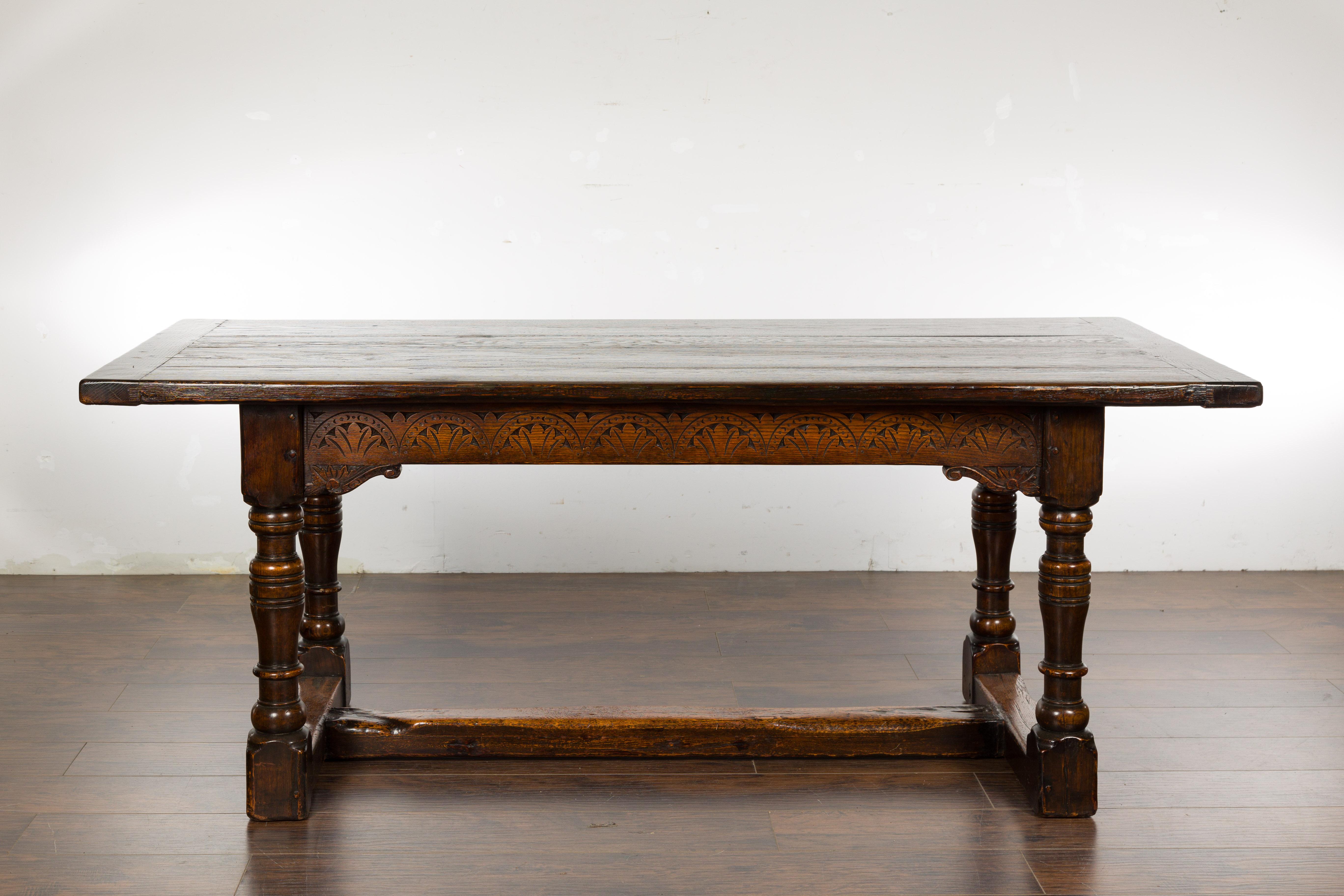 An English oak table from the 19th century with carved apron, turned legs and H-Form cross stretcher. An English oak table from the 19th century, exuding a distinct charm that captures the essence of English craftsmanship. Constructed with robust