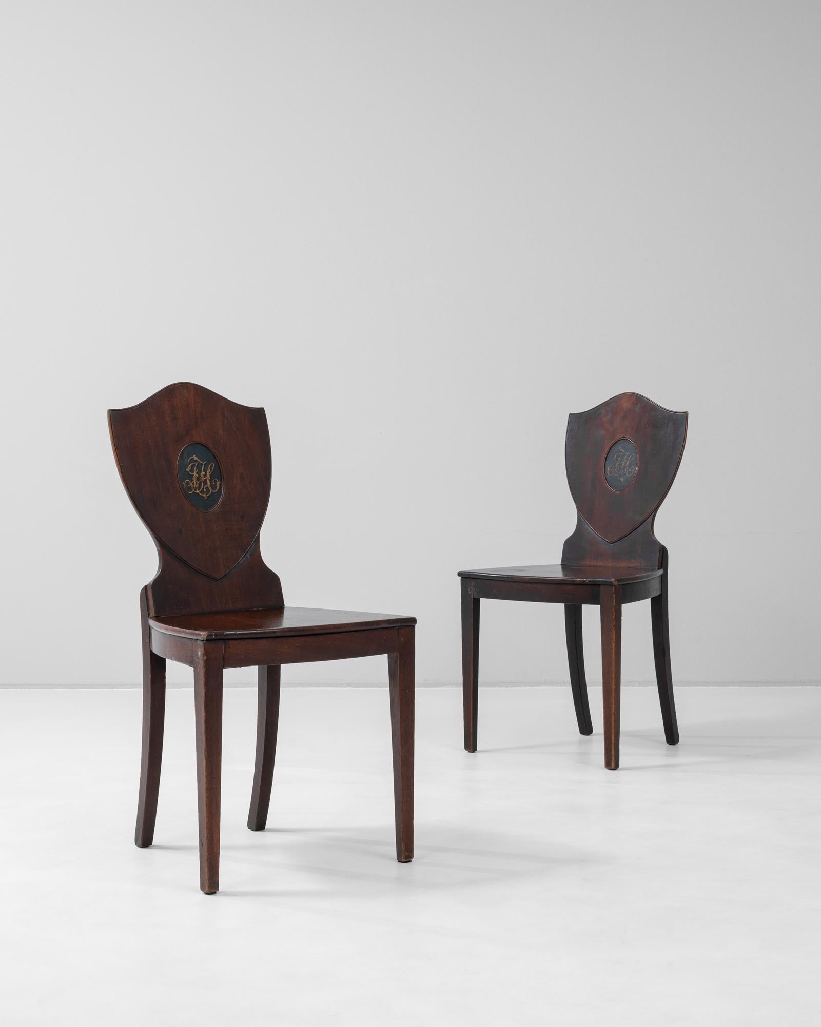 This 19th century English pair of chairs features a distinctive shield-shaped backrest, each adorned with a painted medallion monogram, adding a personalized and historic touch. Crafted from richly patinated wood, the chairs showcase clean, tapering